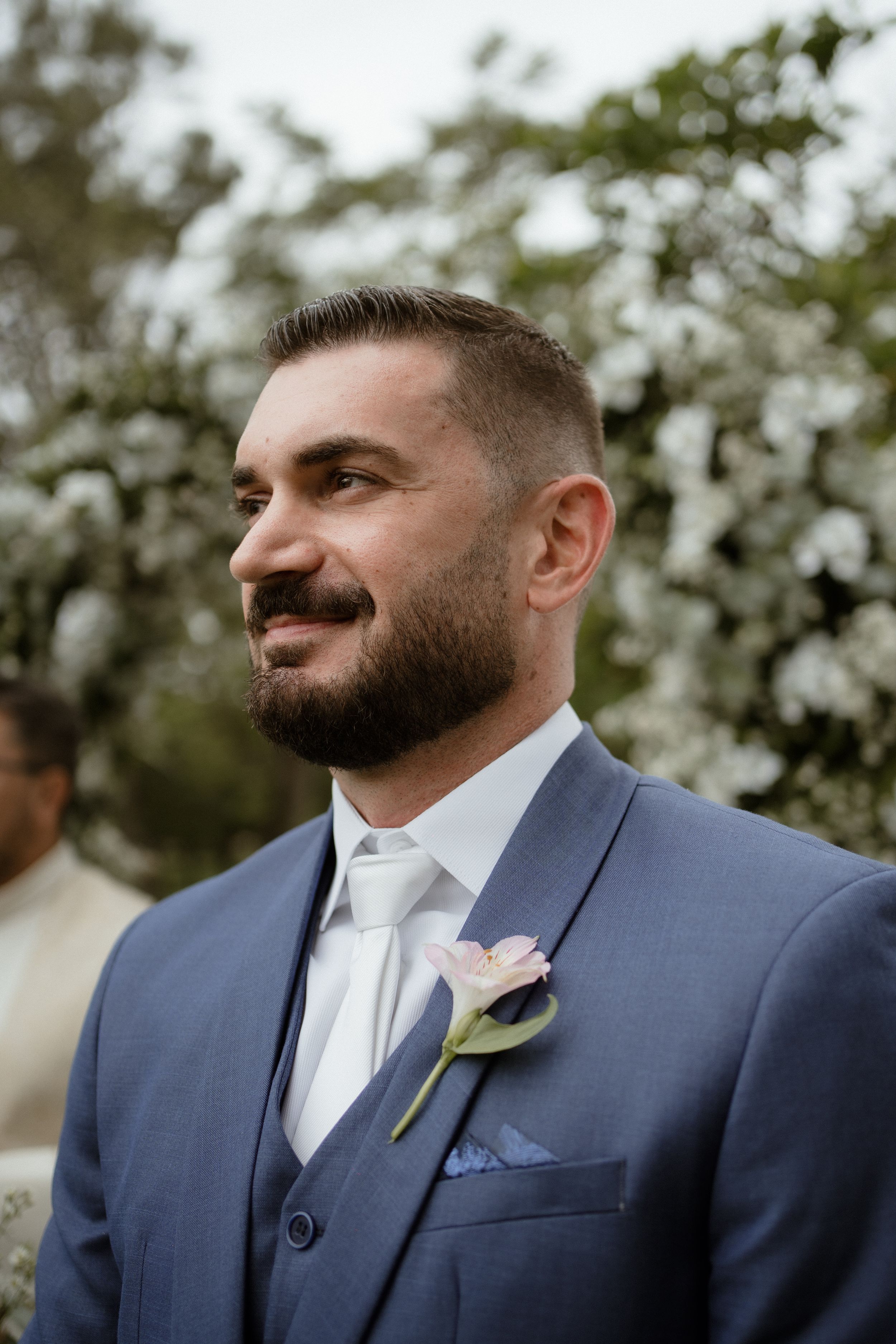 Groom in a blue suit with a white tie and pink boutonniere stands in front of blooming white flowers.