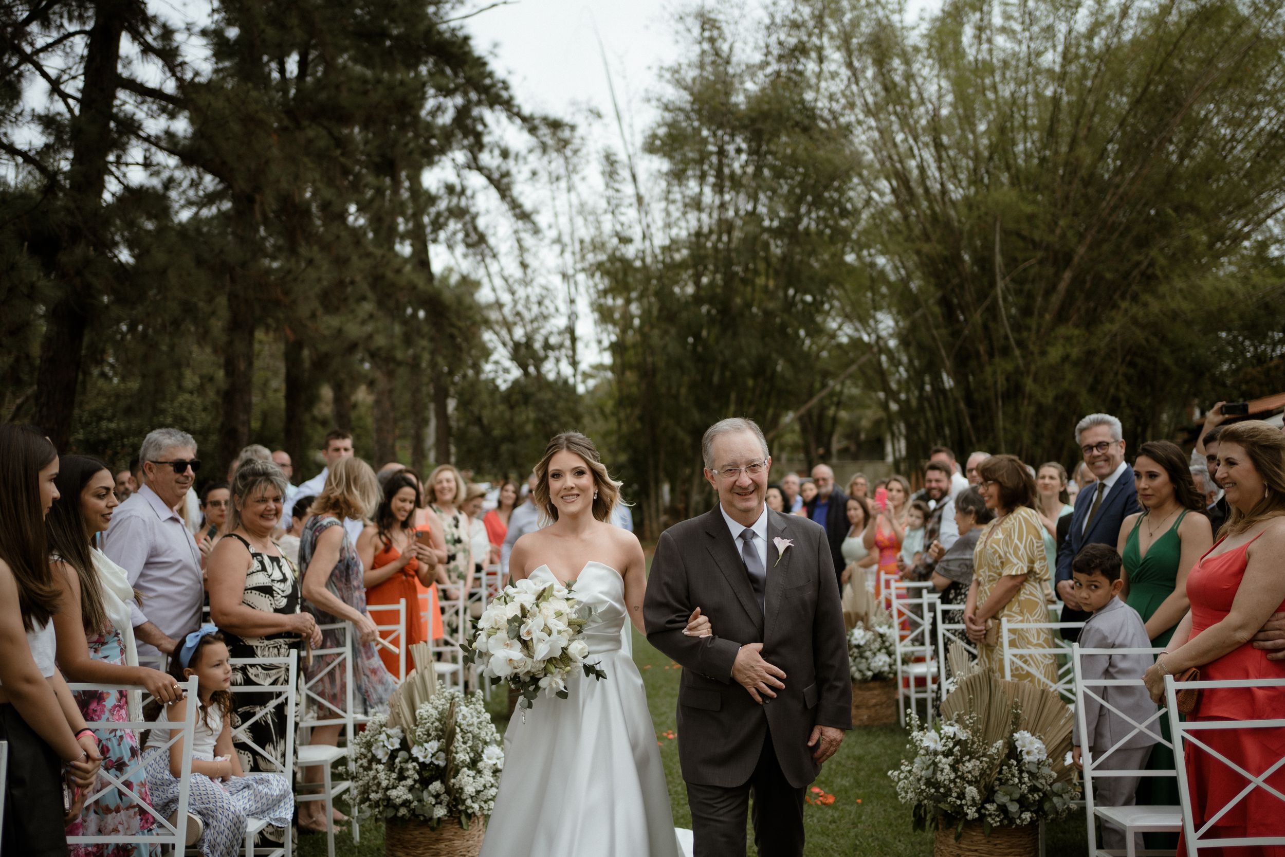 Wedding guests seated in white chairs for an outdoor ceremony under tall trees.
