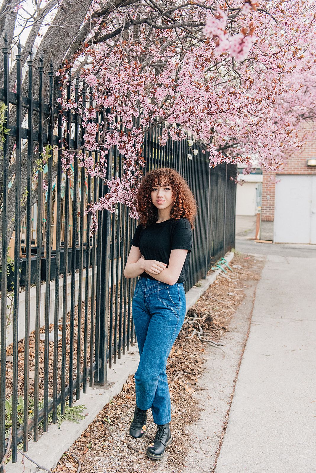 Portrait against pink cherry blossoms and iron fence in casual outfit with curly hair.