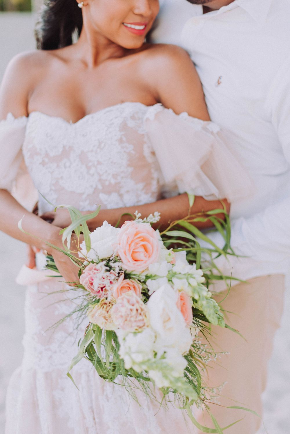 A close-up of a bridal bouquet with blush pink and white roses against a white lace wedding dress.