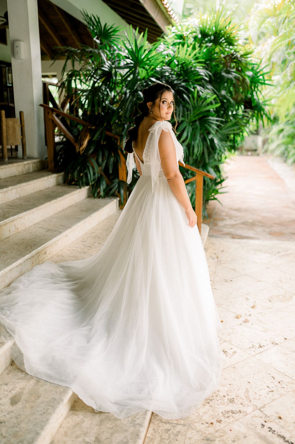 Bride in flowing white wedding gown poses against tropical garden backdrop.