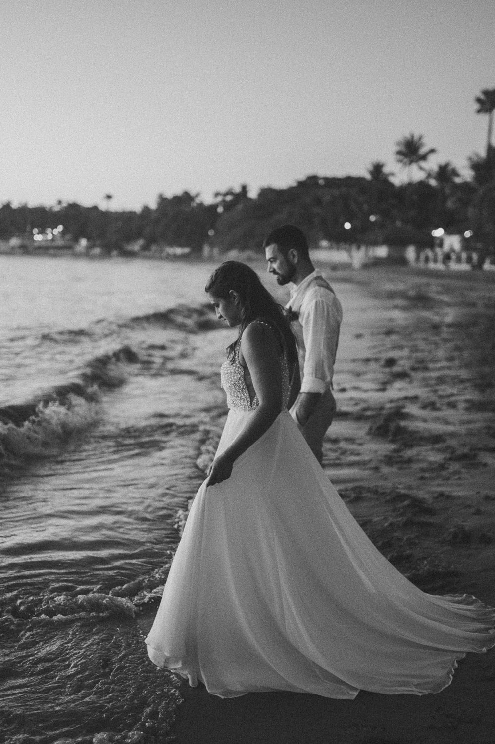 A couple walks along the shoreline at sunset with flowing white wedding dress.