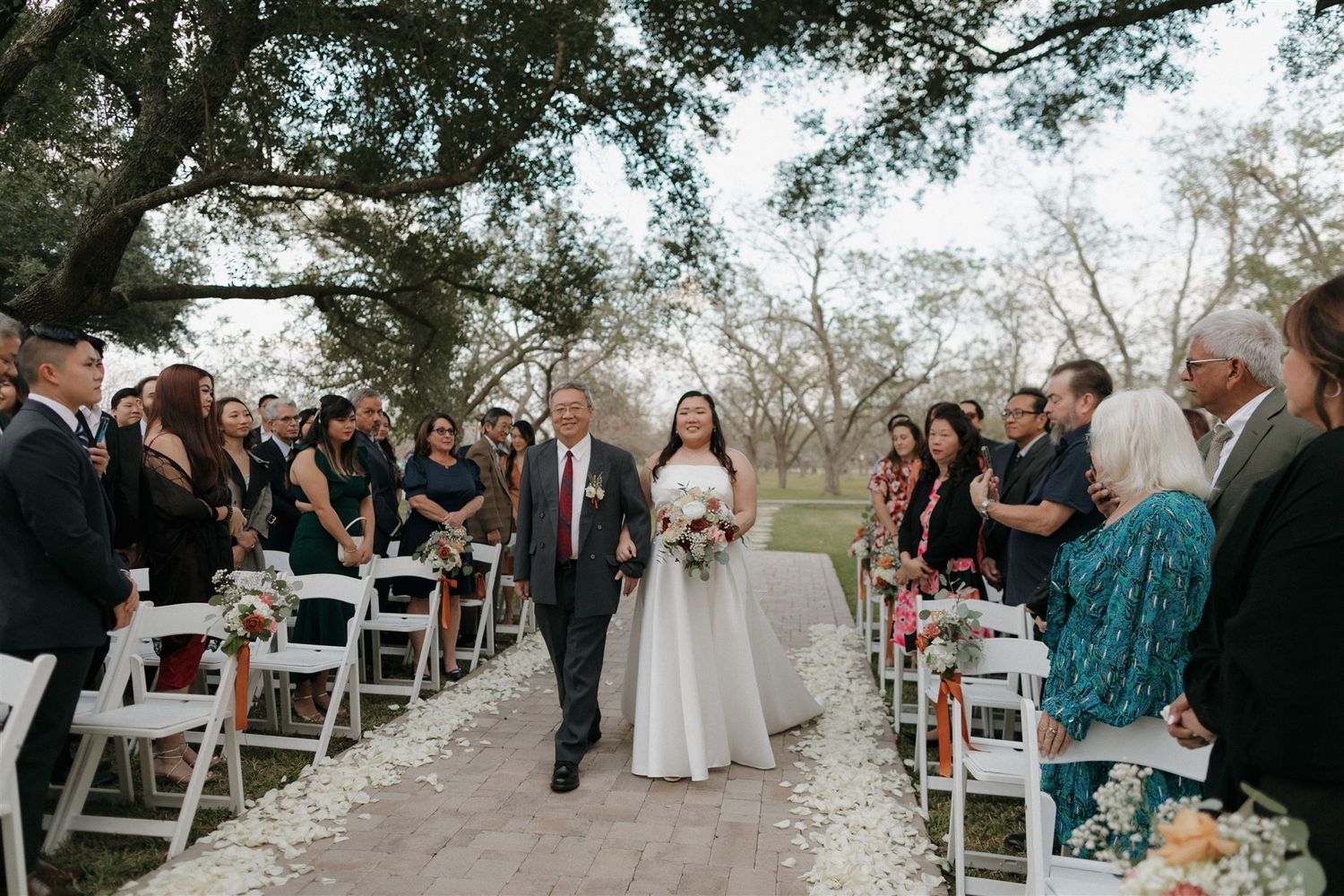 Guests watch as the wedding party proceeds down an outdoor aisle strewn with white petals.