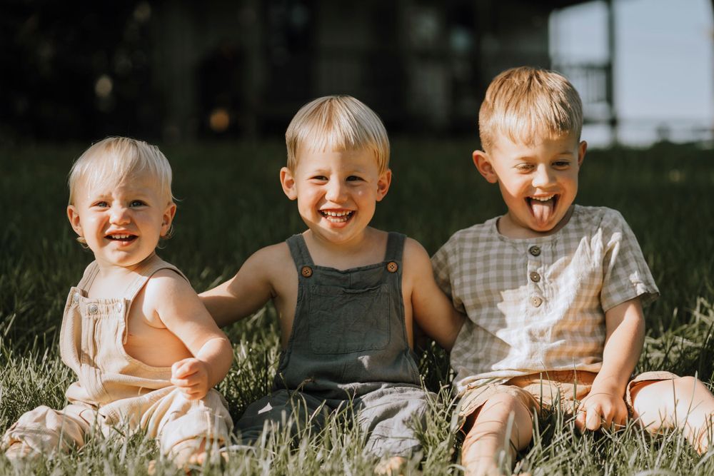 Three children sit together in grass, smiling and laughing on a sunny day.