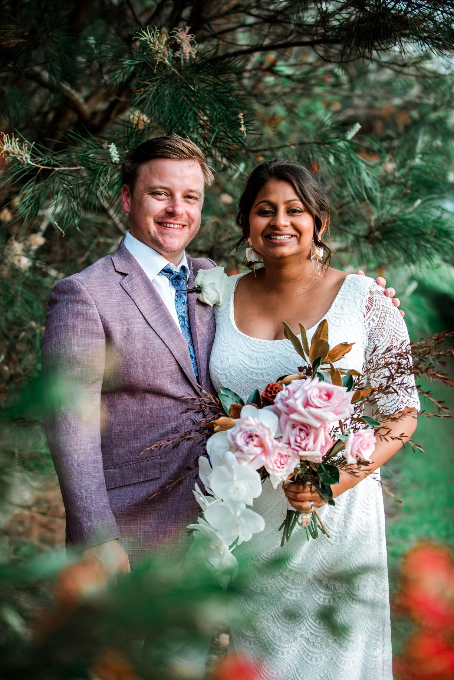 A couple in wedding attire poses among pine trees holding a pink and white floral bouquet.