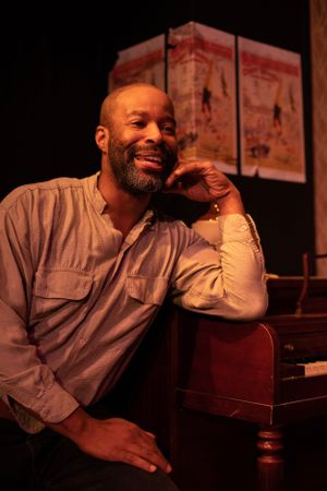 A performer in casual attire poses beside a wooden piano on stage.