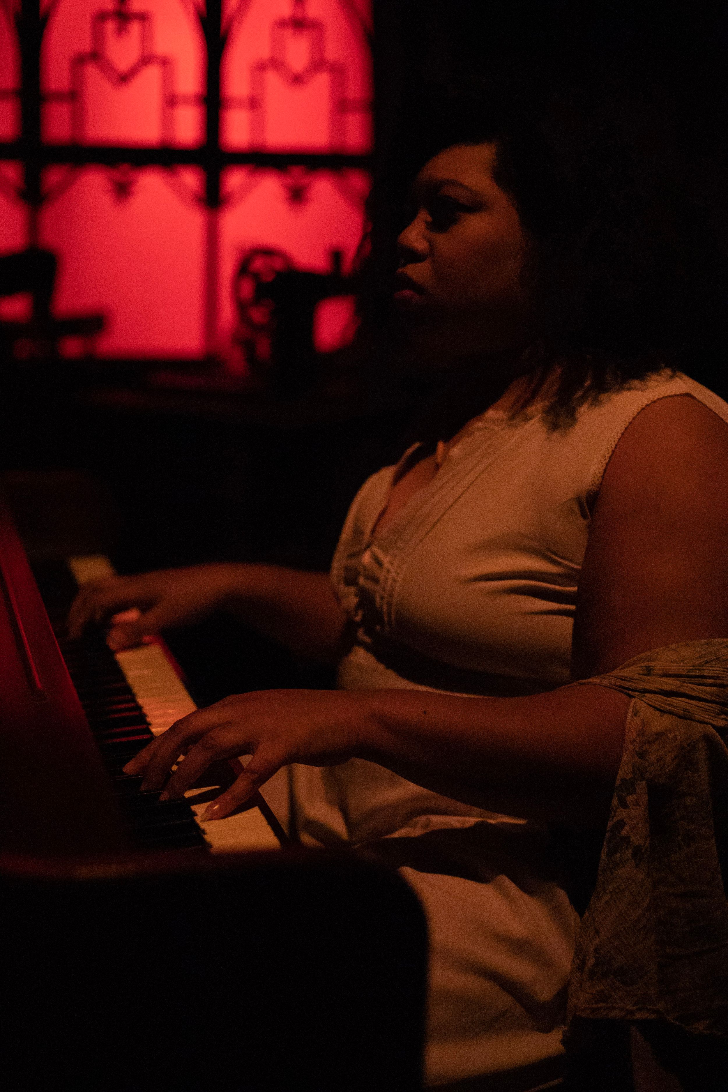A performer plays piano under dramatic red lighting with gothic windows in background.