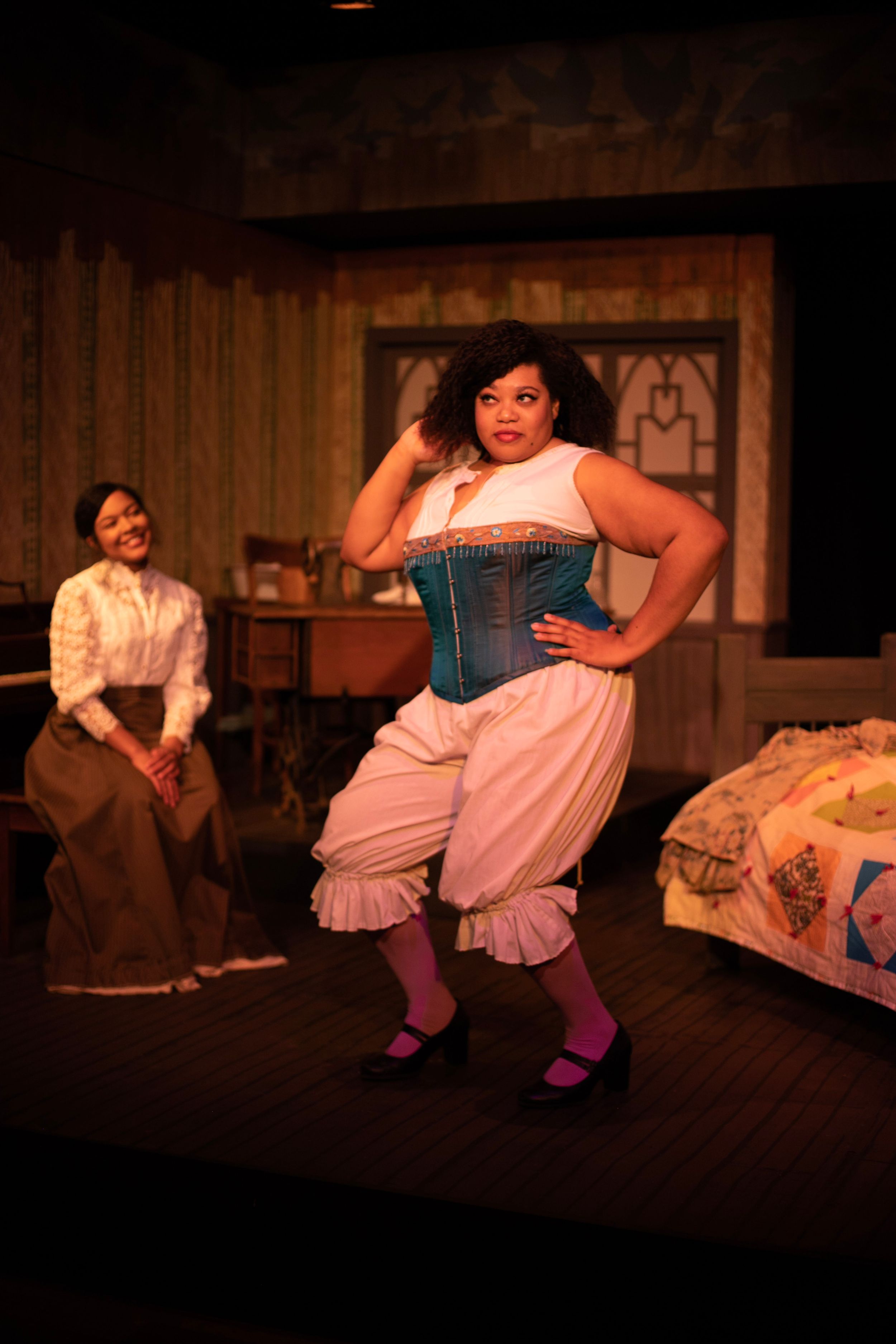 A performer in a teal corset and bloomers strikes a pose while another watches from behind.