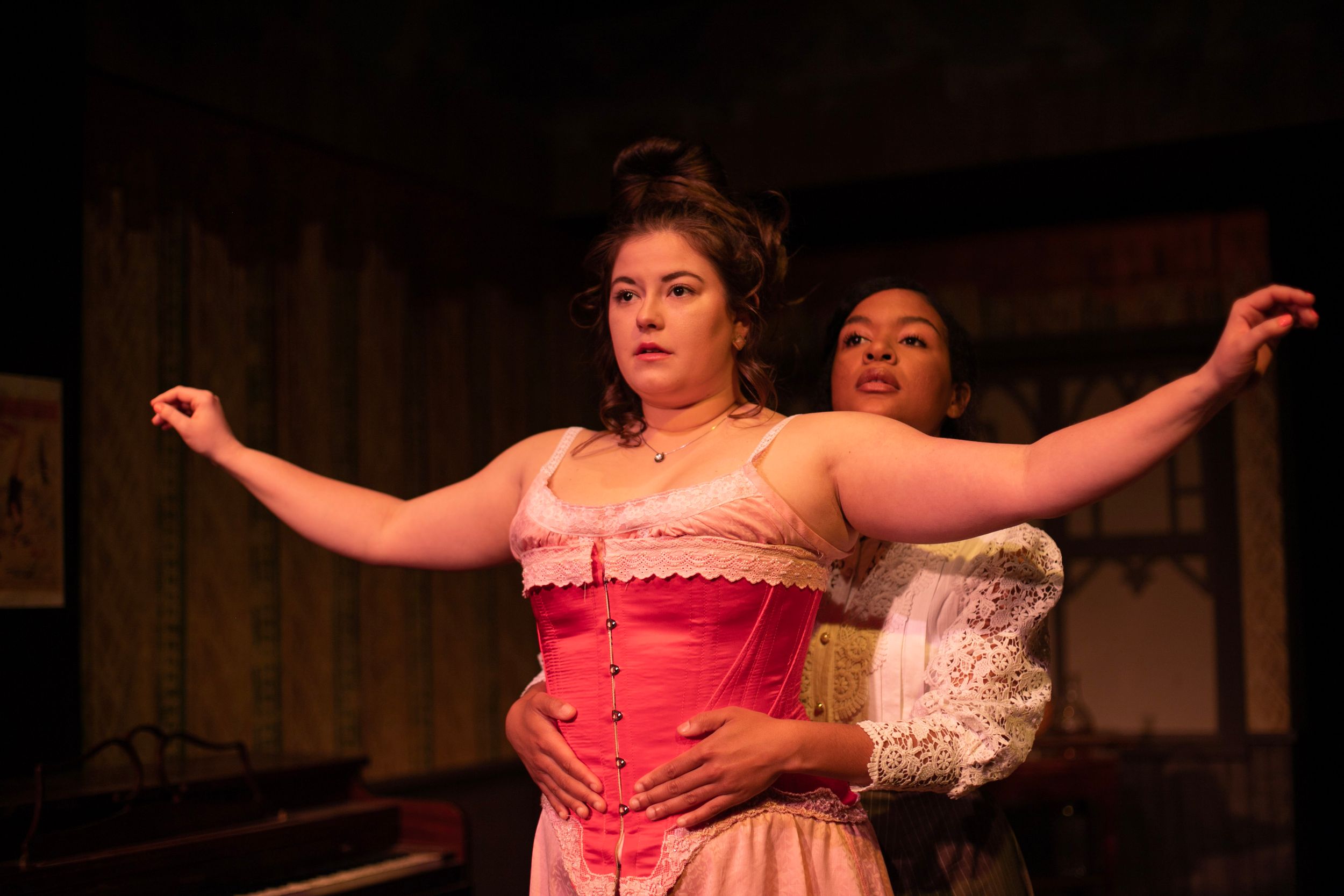 Two performers in period costumes embrace during a theatrical scene on stage.