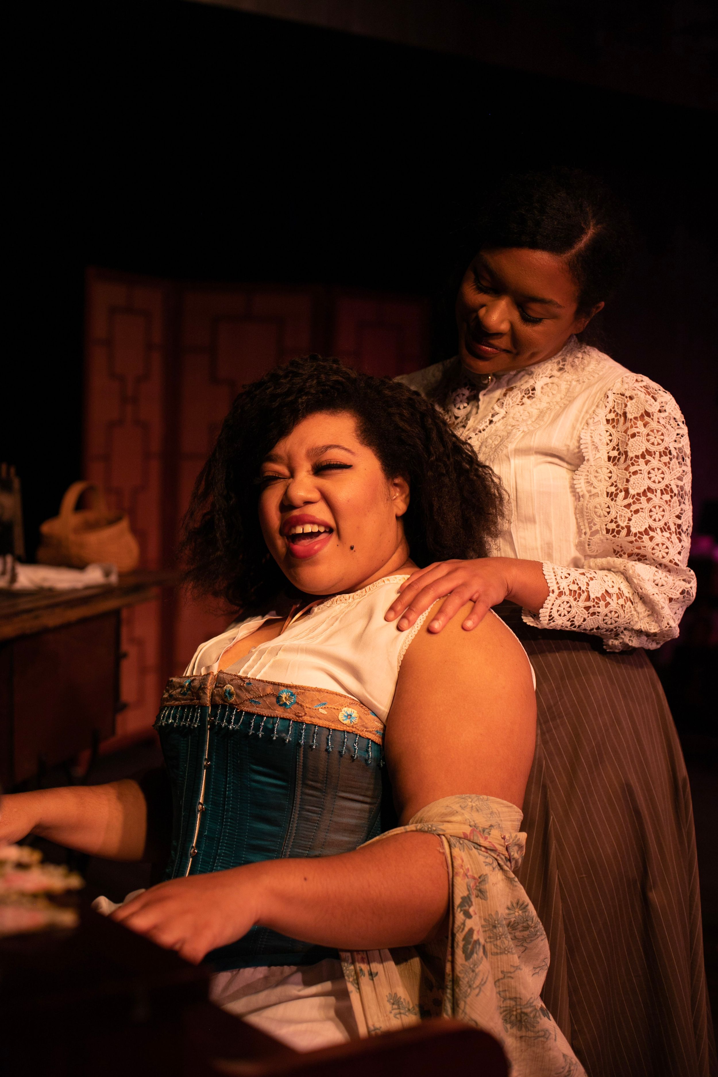 Two performers share a dramatic moment in period costumes with theatrical lighting.