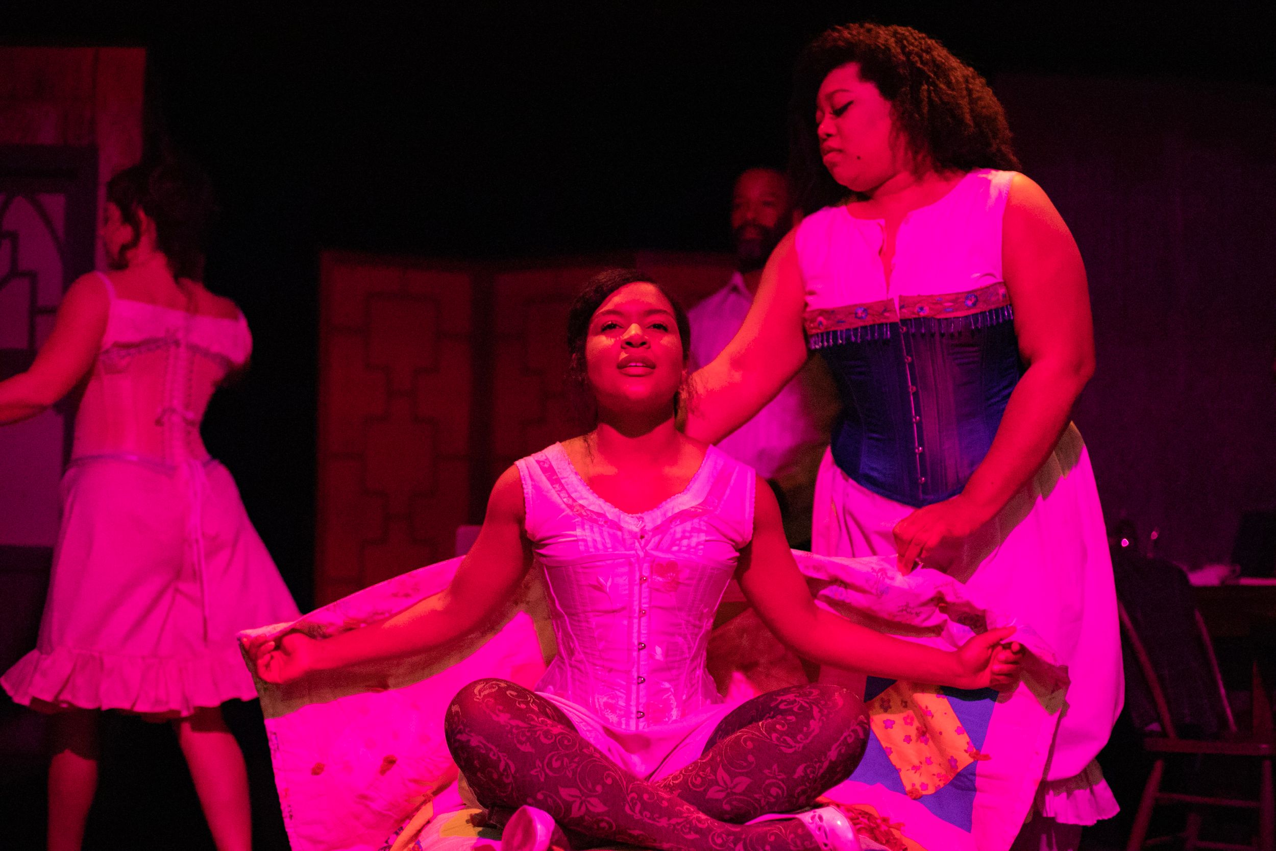 Performers in pink corsets and dresses pose dramatically under theatrical lighting.