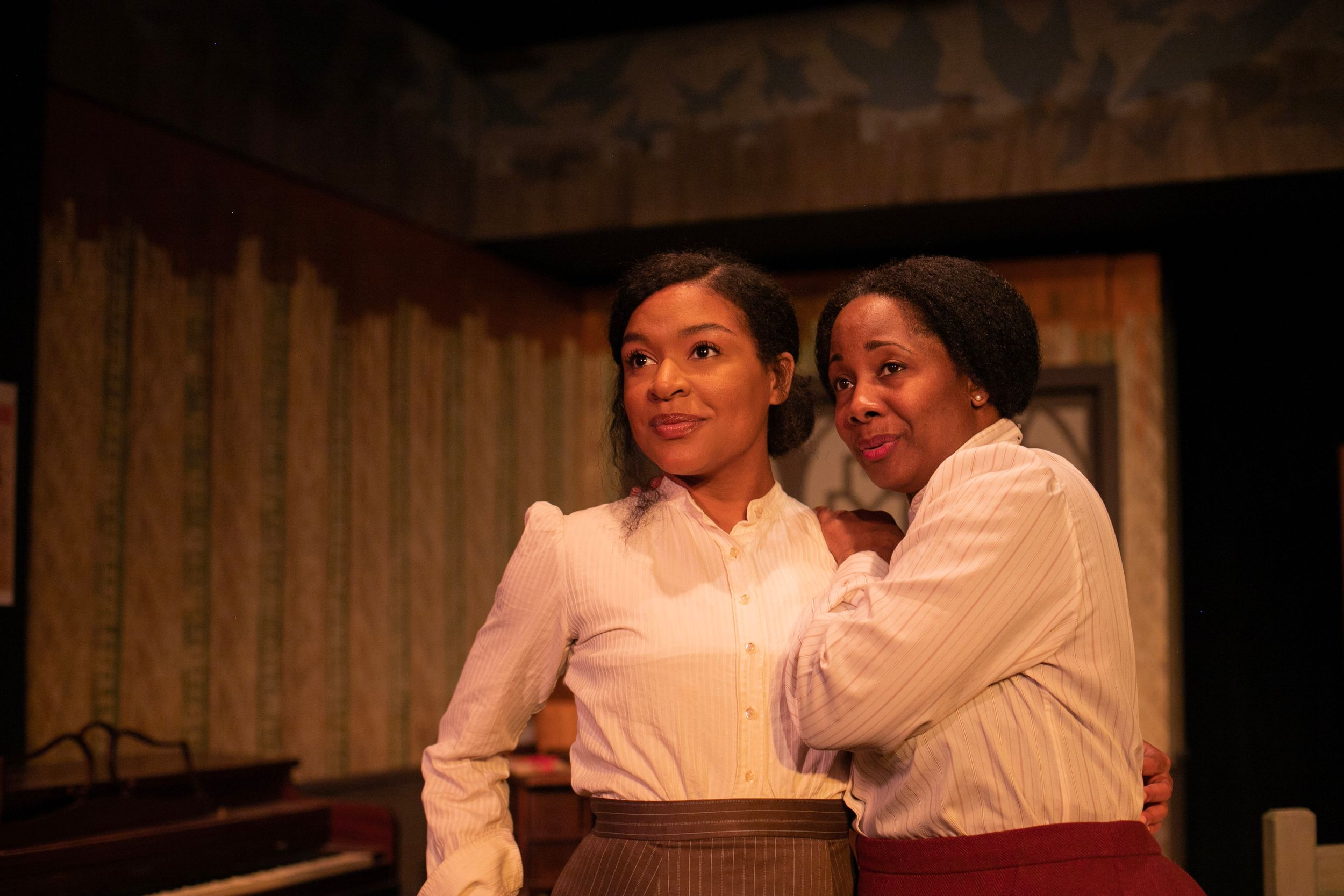 Two performers share an intimate moment in period costumes on a dimly lit stage.