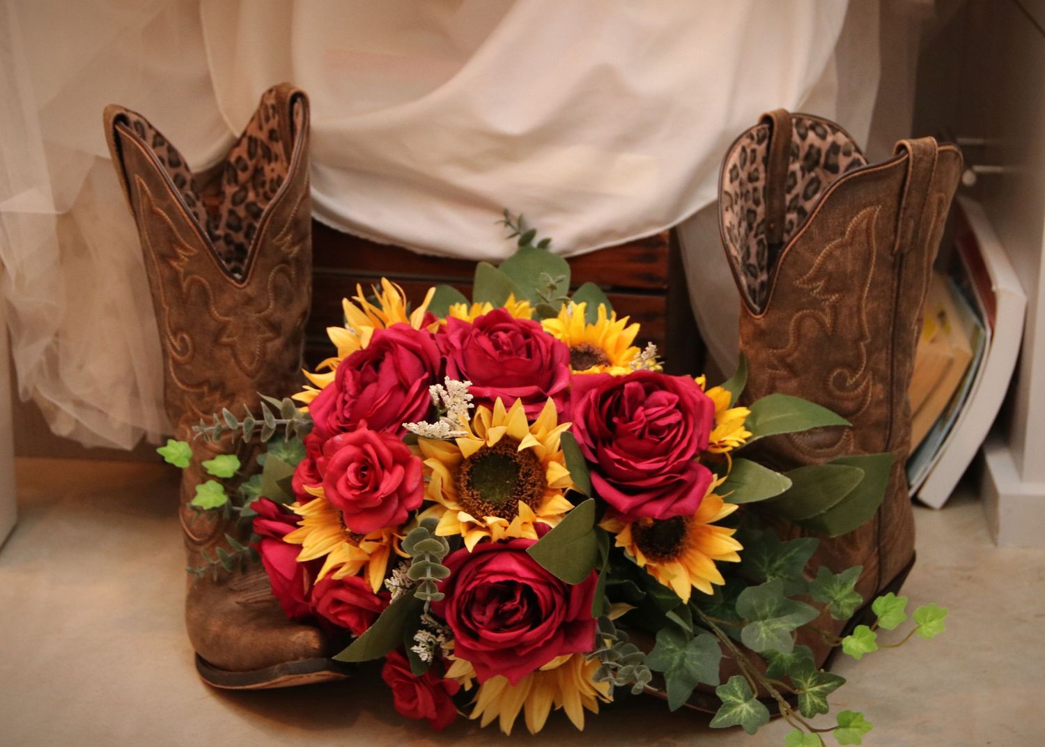 A rustic wedding bouquet with red roses and sunflowers placed between brown cowboy boots.