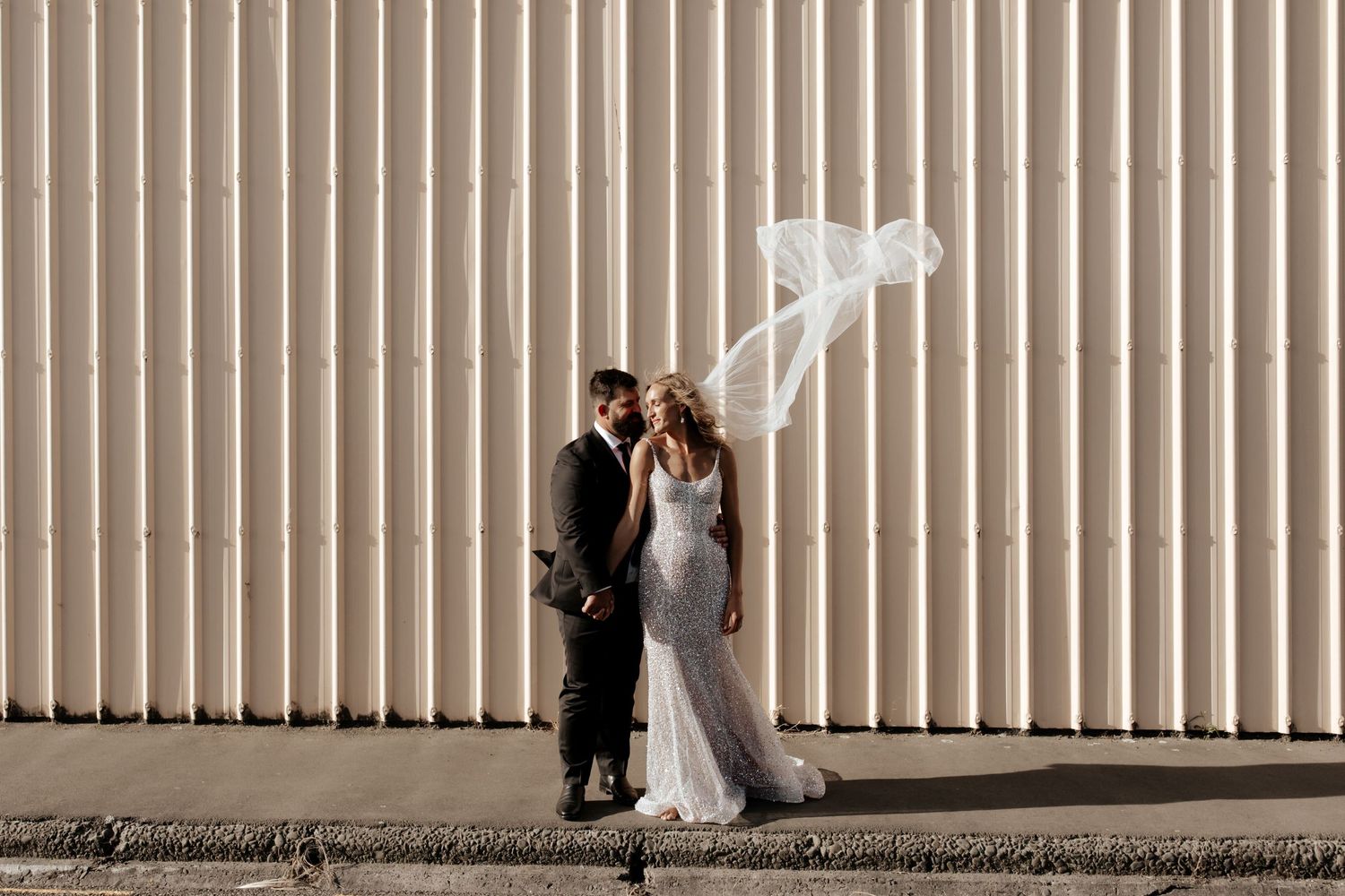 Wedding couple stands against industrial wall as bride's veil billows dramatically in the wind.