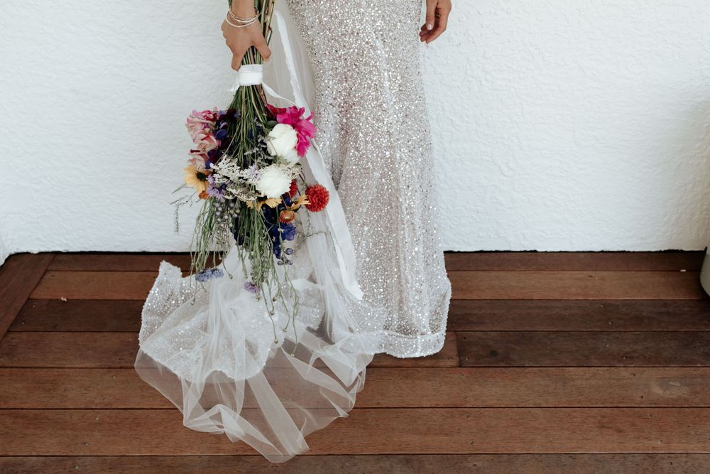 Detail shot of sequined wedding dress hem and veil with colorful floral bouquet on wooden floor.