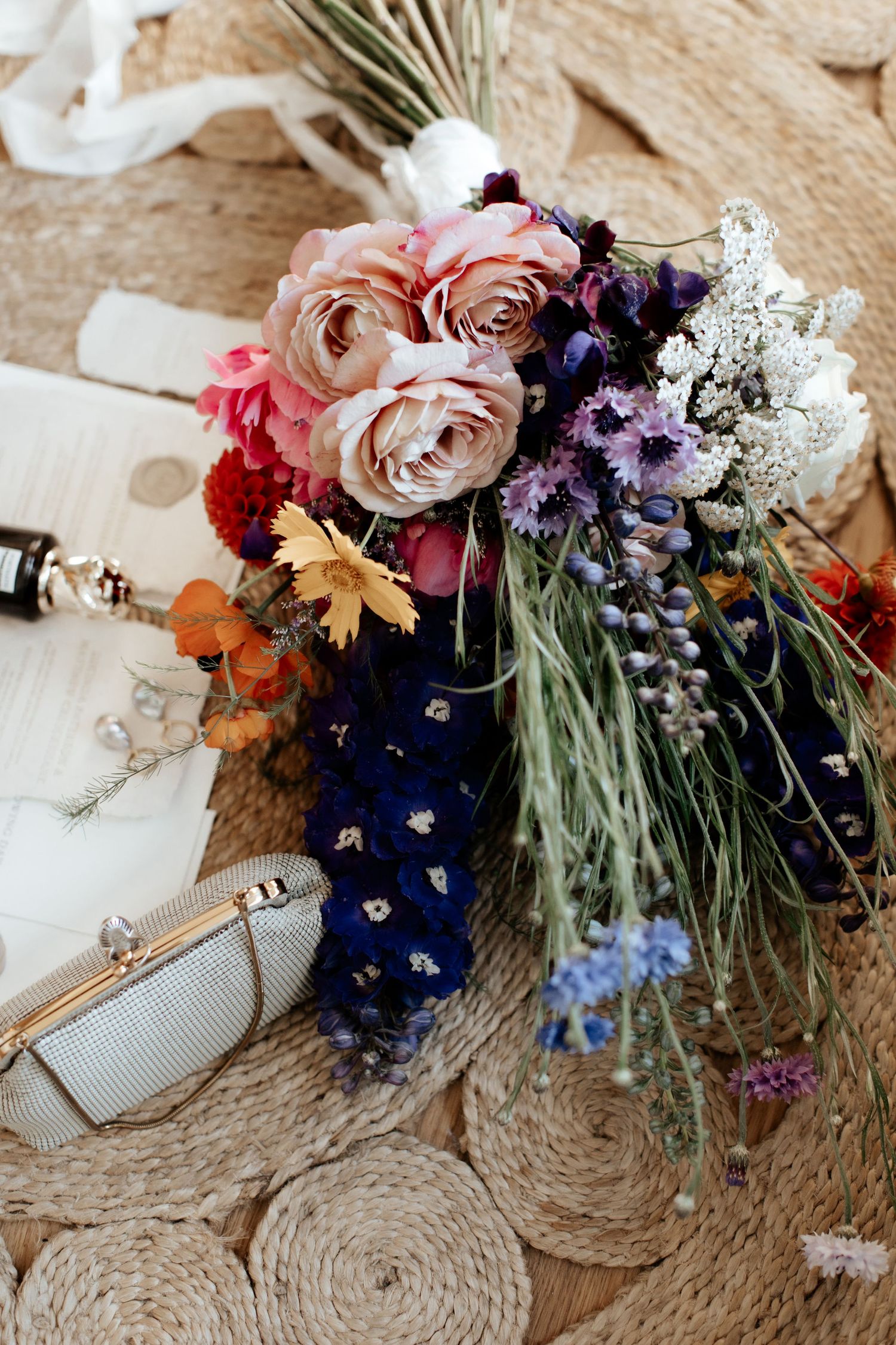 Wedding bouquet with pink roses and purple flowers alongside silver clutch on woven surface.