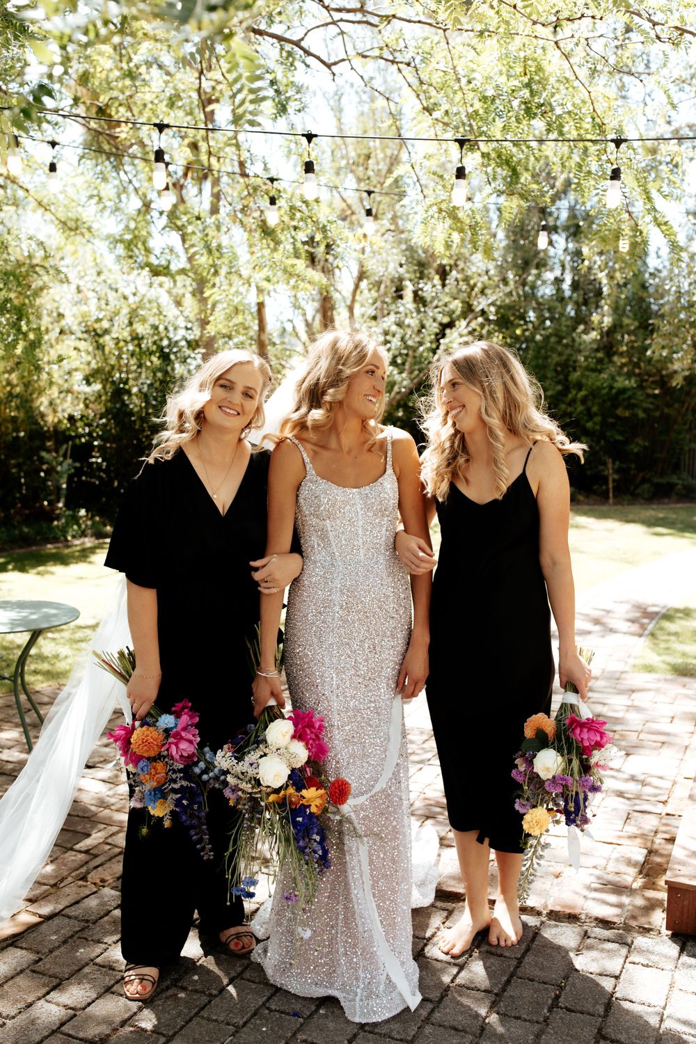 Wedding party poses on brick patio with bride in sparkly gown flanked by attendants in black dresses.