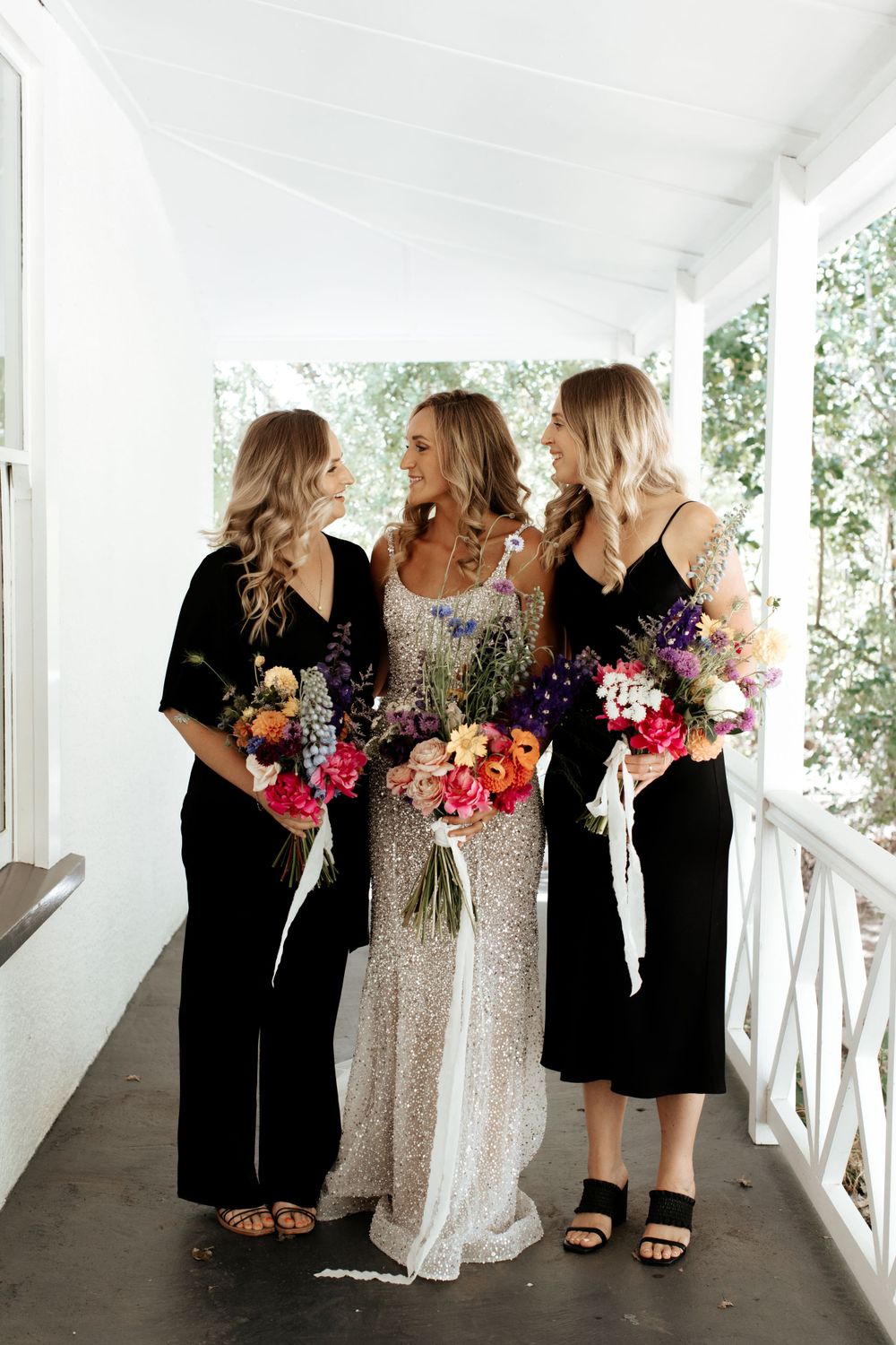 Three women on a white porch holding colorful bouquets, with two in black dresses and one in a sparkly gown.