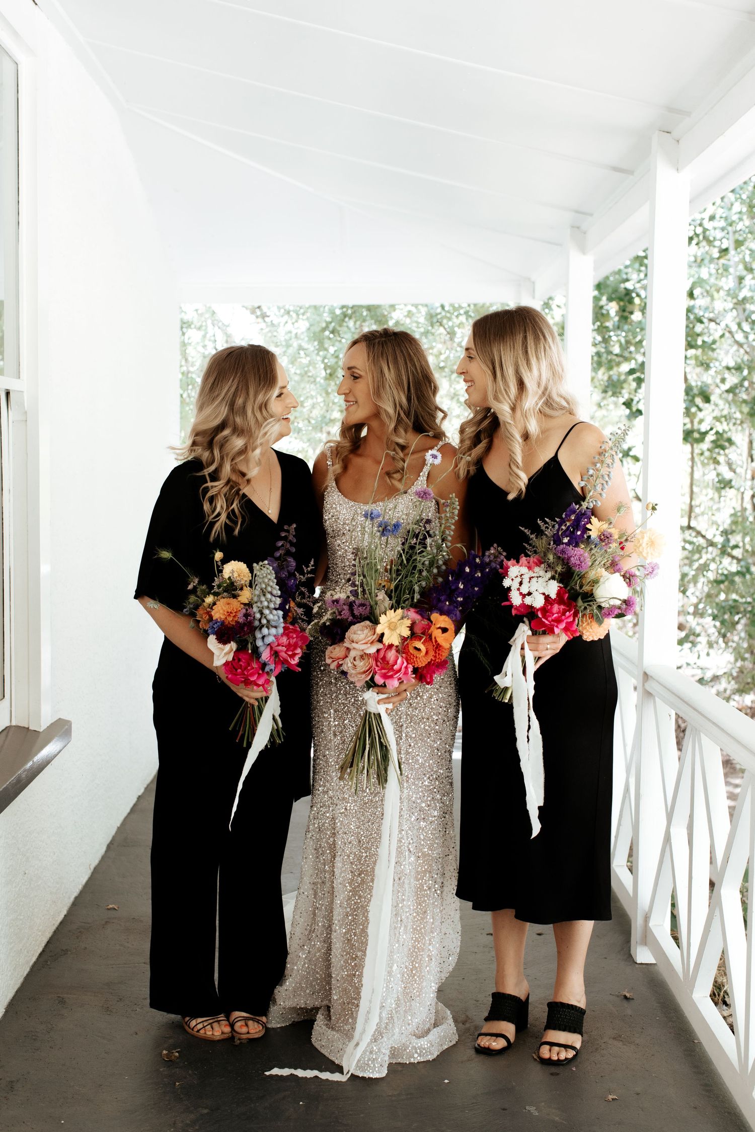 Three women on a white porch holding colorful bouquets, with two in black dresses and one in a sparkly gown.