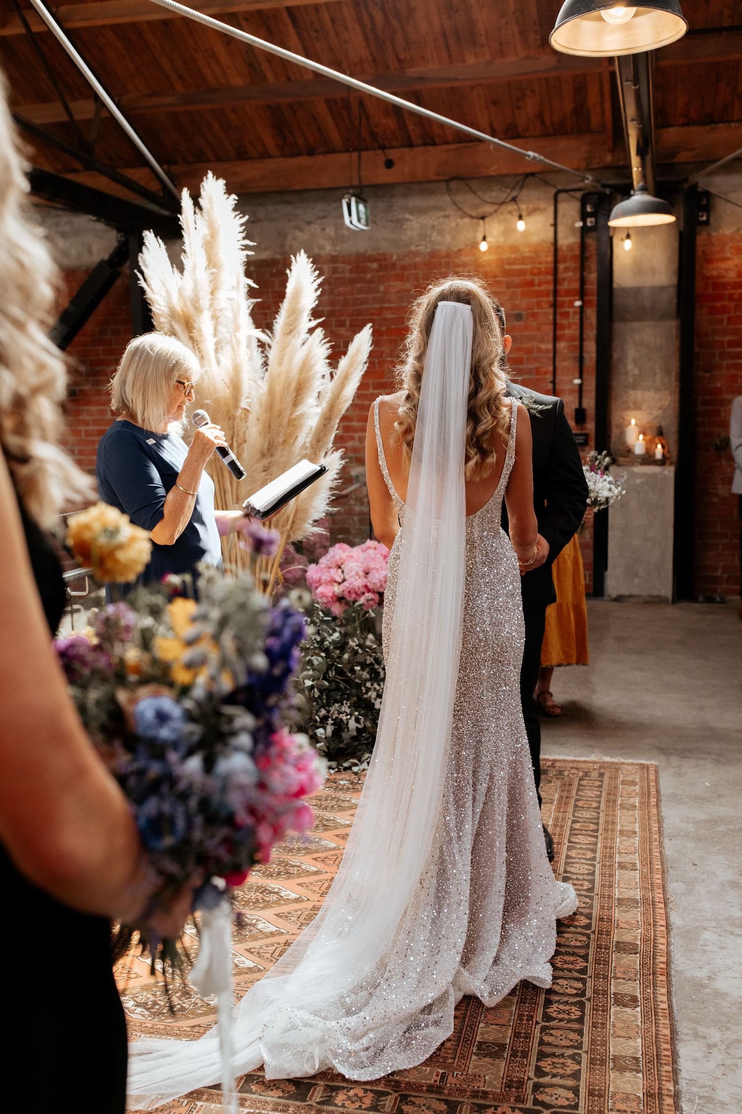 Bride in sparkling gown with flowing veil stands before pampas grass decorations in industrial venue.