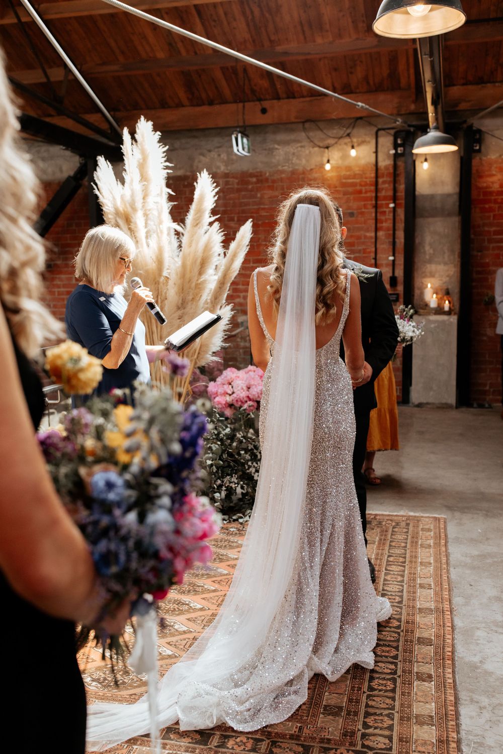 Bride in sparkling gown with flowing veil stands before pampas grass decorations in industrial venue.