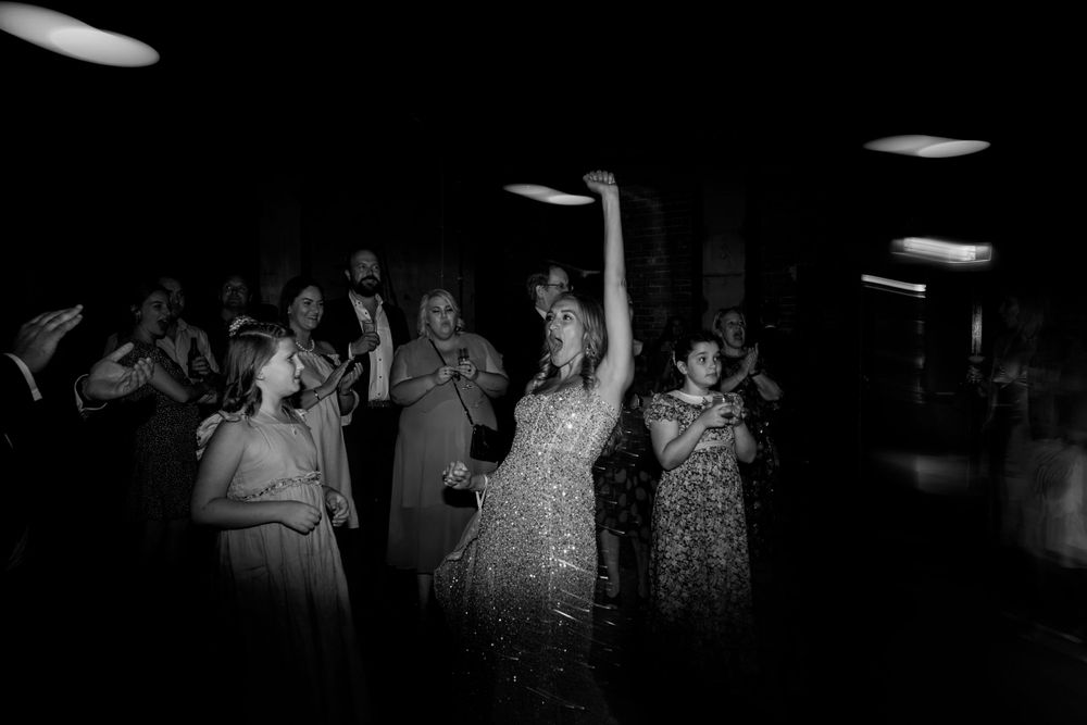 Wedding guests dancing and celebrating in a dimly lit venue with overhead lighting.