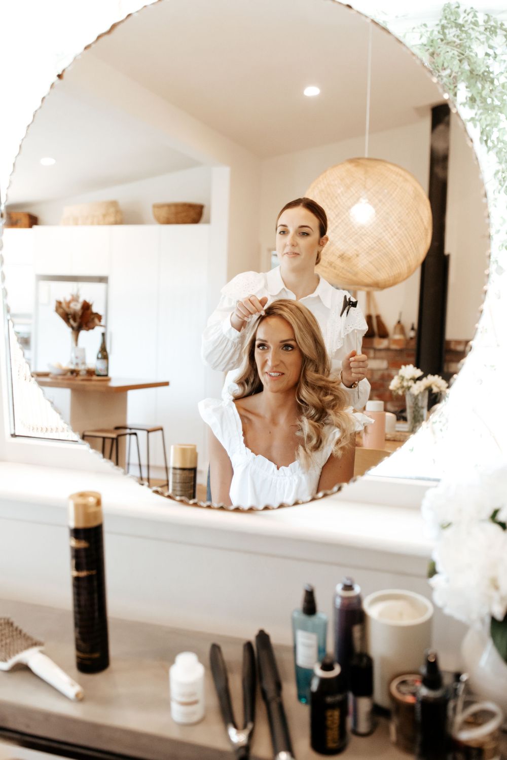 Getting ready scene reflected in round mirror with beauty products and pendant lighting.