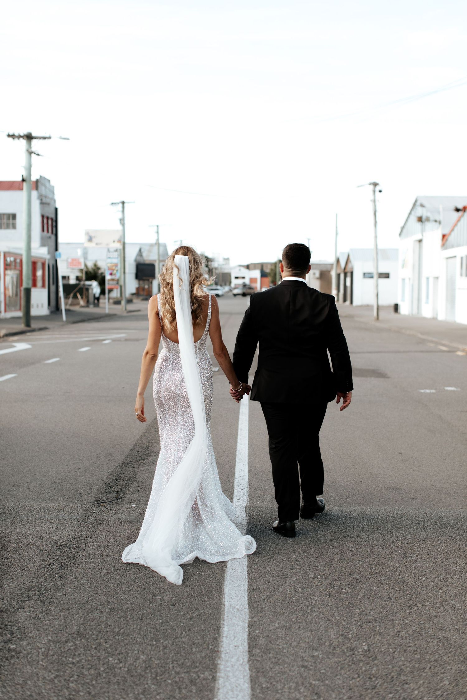 Couple walks hand in hand down empty street with white buildings in background.