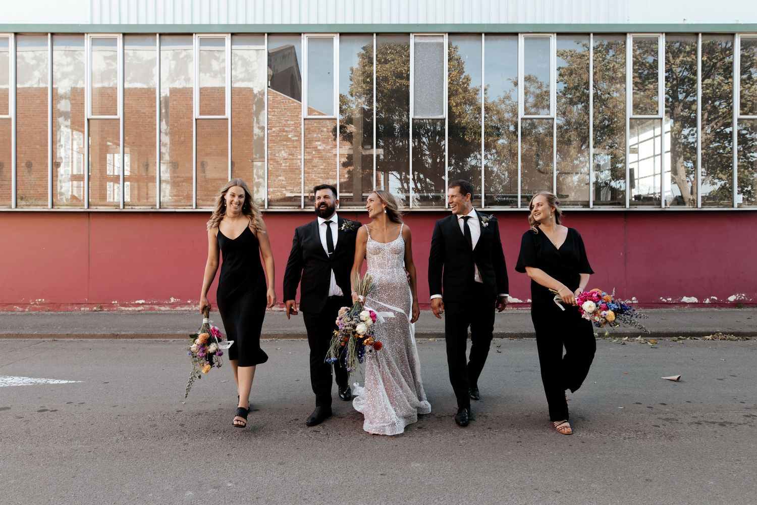 Wedding party in black and white attire walking together in front of a red industrial wall.