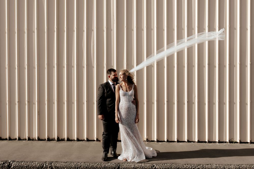 A couple poses against a corrugated metal wall for their wedding portraits with flowing veil.