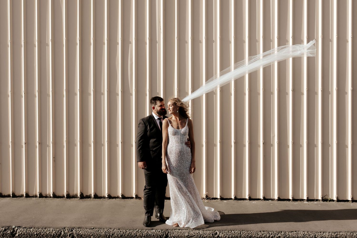A couple poses against a corrugated metal wall for their wedding portraits with flowing veil.
