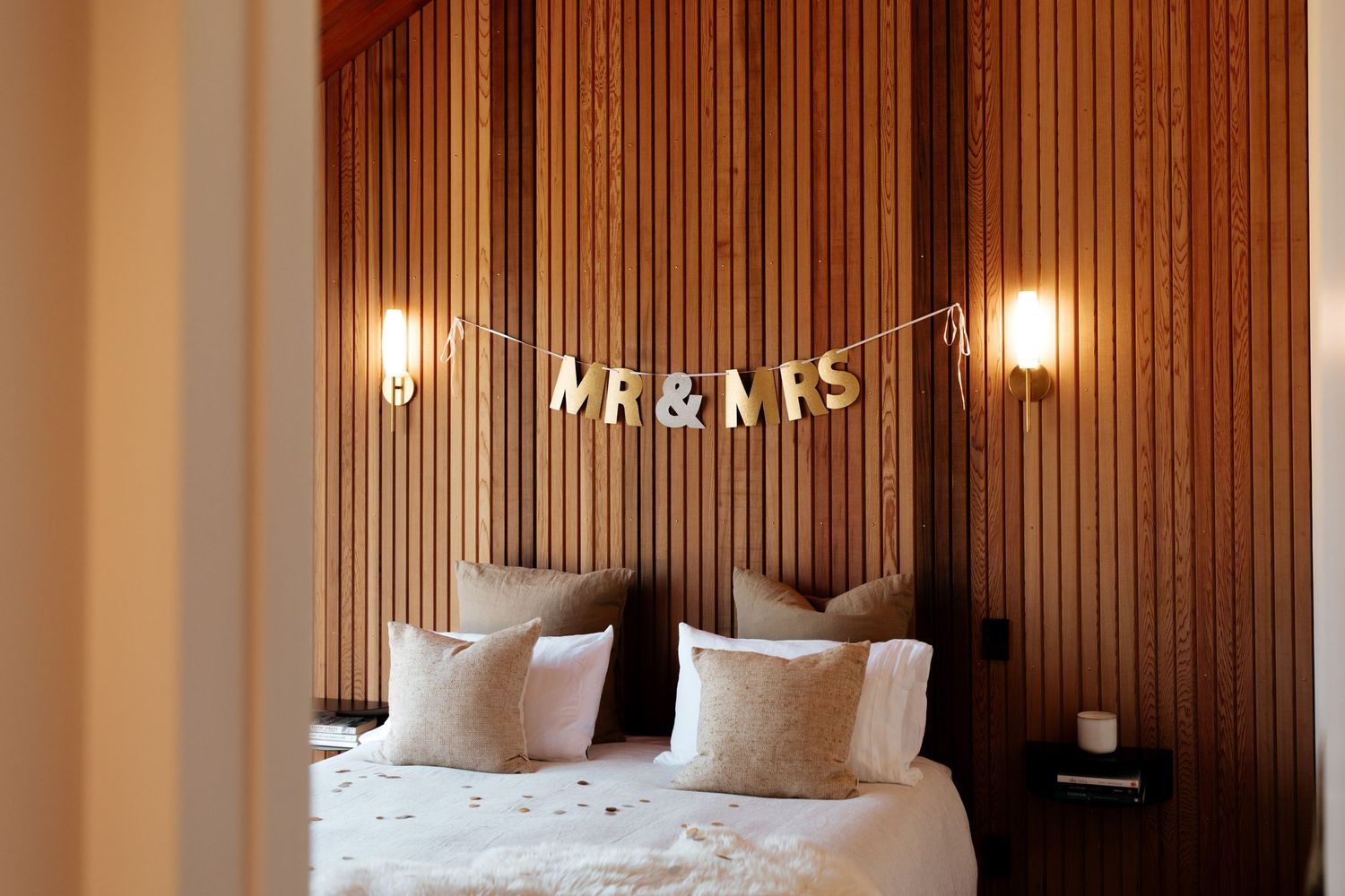 Wooden paneled bedroom wall with 'MR & MRS' text banner and wall sconces above a white bed.