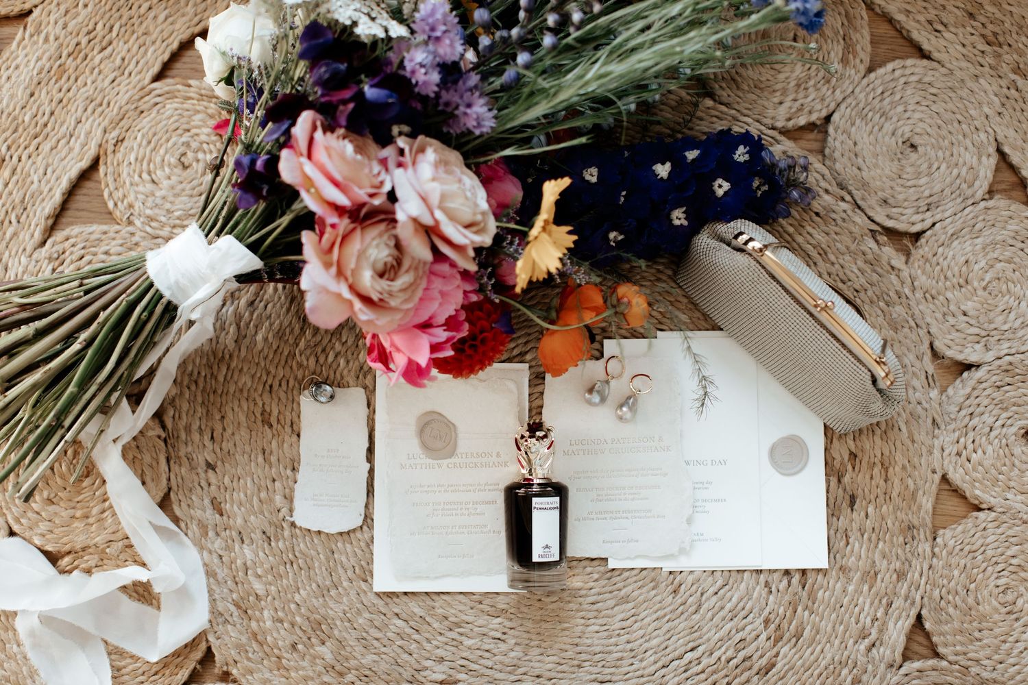 Wedding stationery and perfume bottle arranged with floral bouquet on circular jute rug.