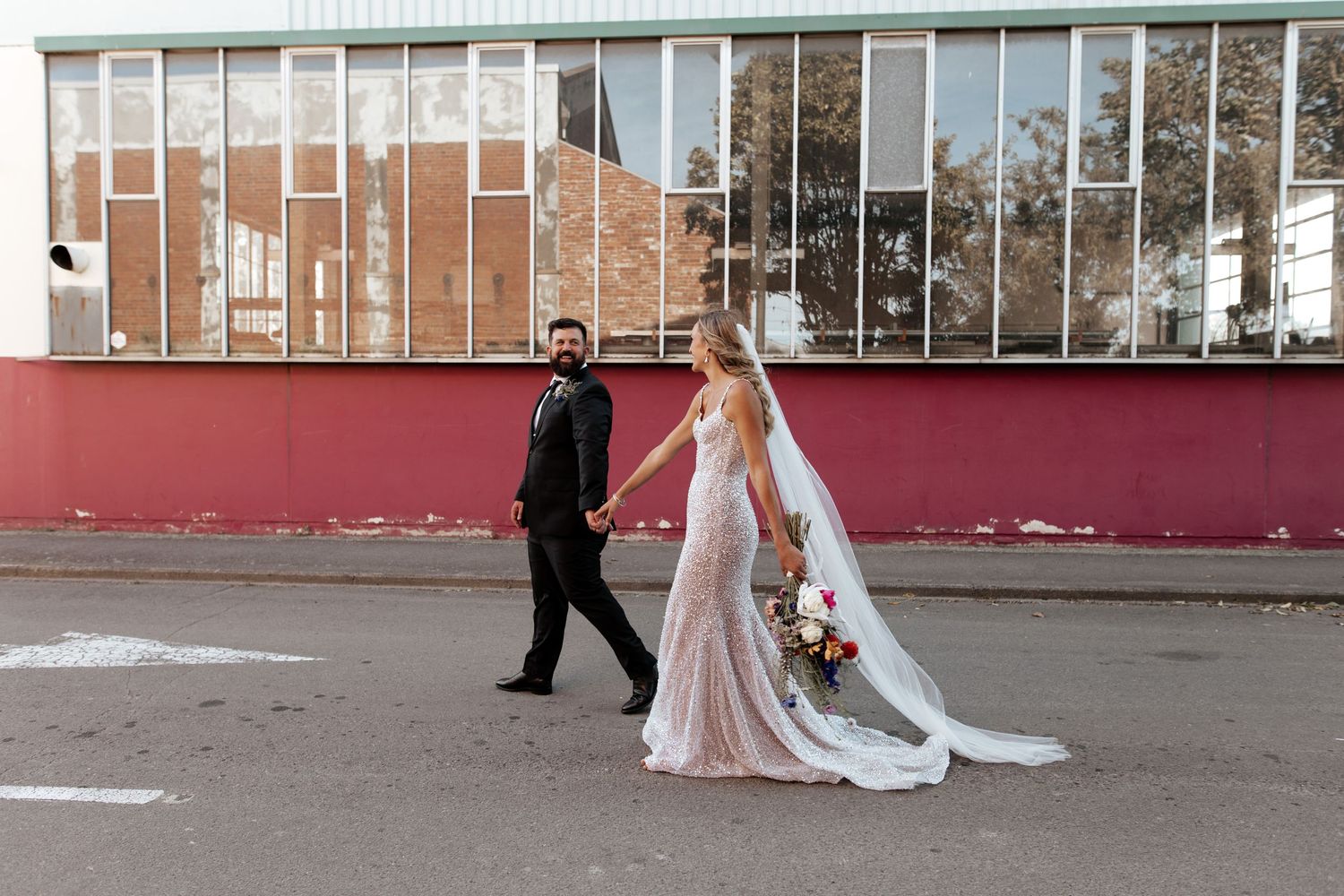 A couple walks hand in hand in wedding attire past a modern architectural facade.