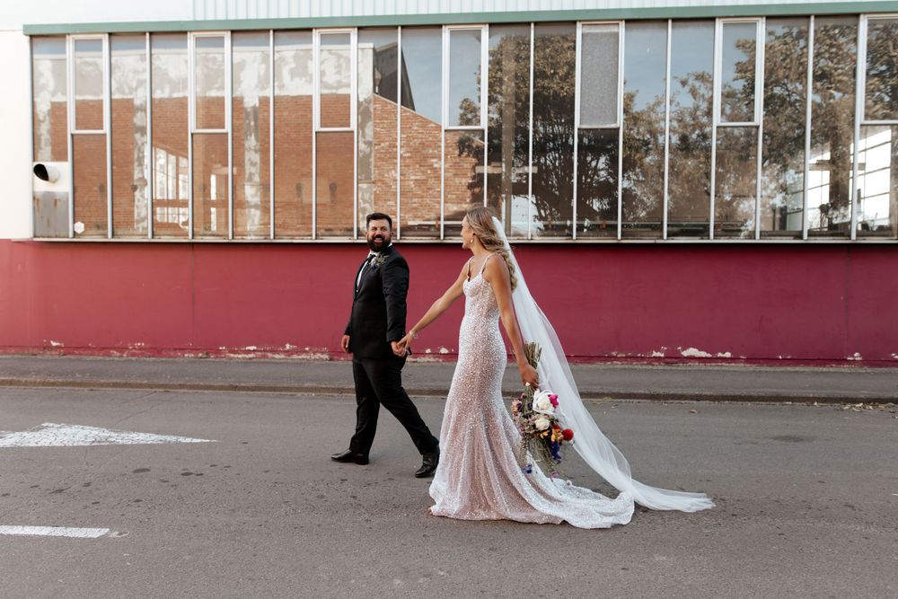 A couple walks hand in hand in wedding attire past a modern architectural facade.