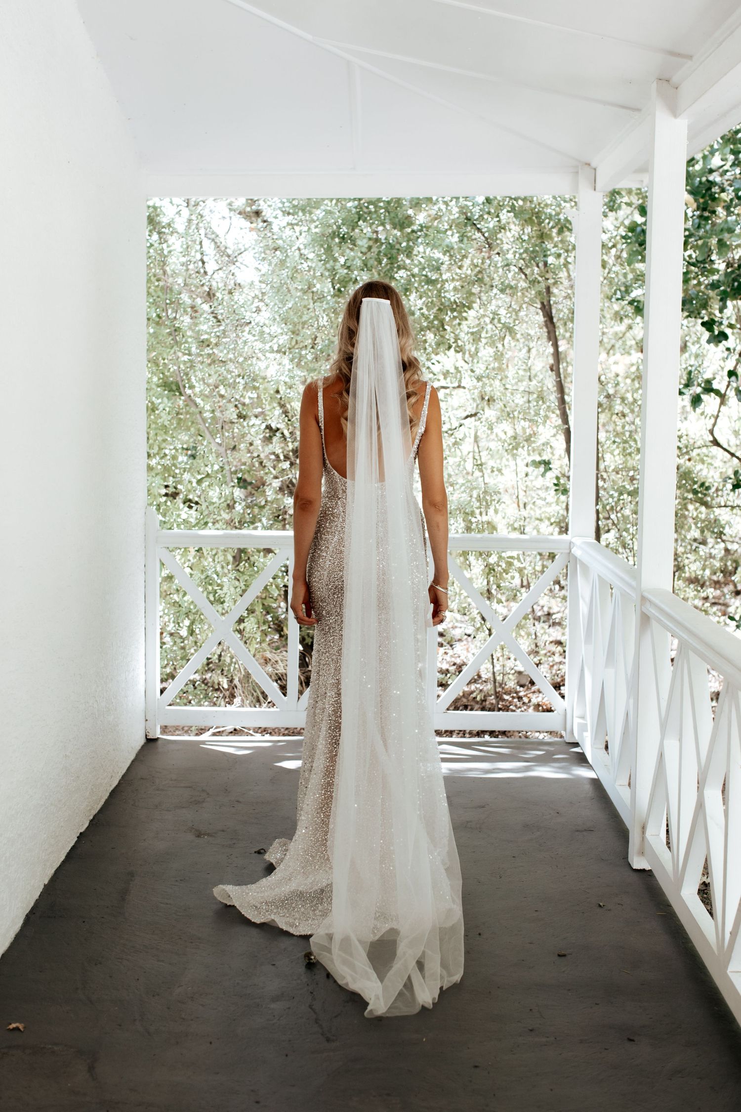 A long white wedding dress with a train photographed from behind on a white porch with natural greenery in background.