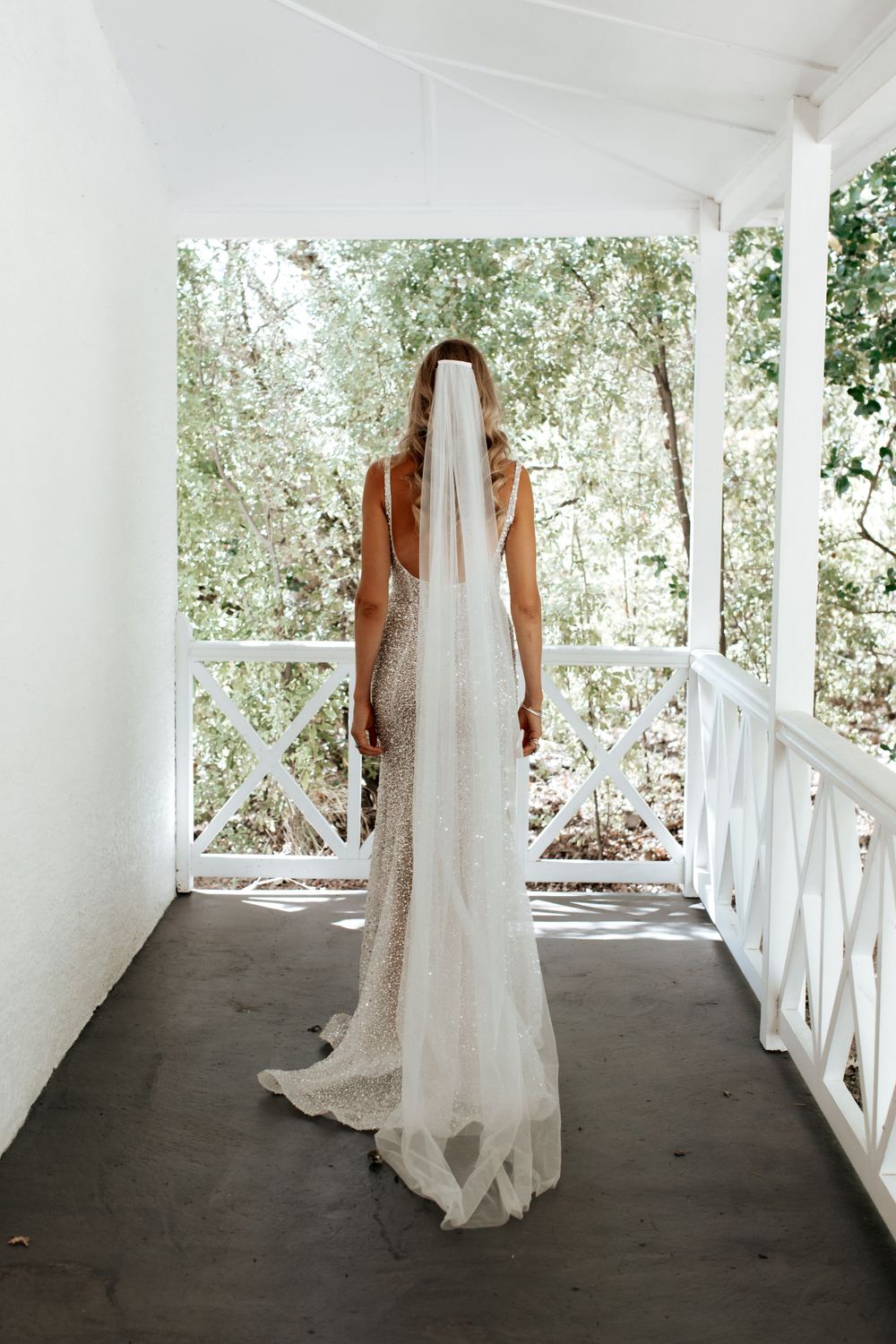 A long white wedding dress with a train photographed from behind on a white porch with natural greenery in background.