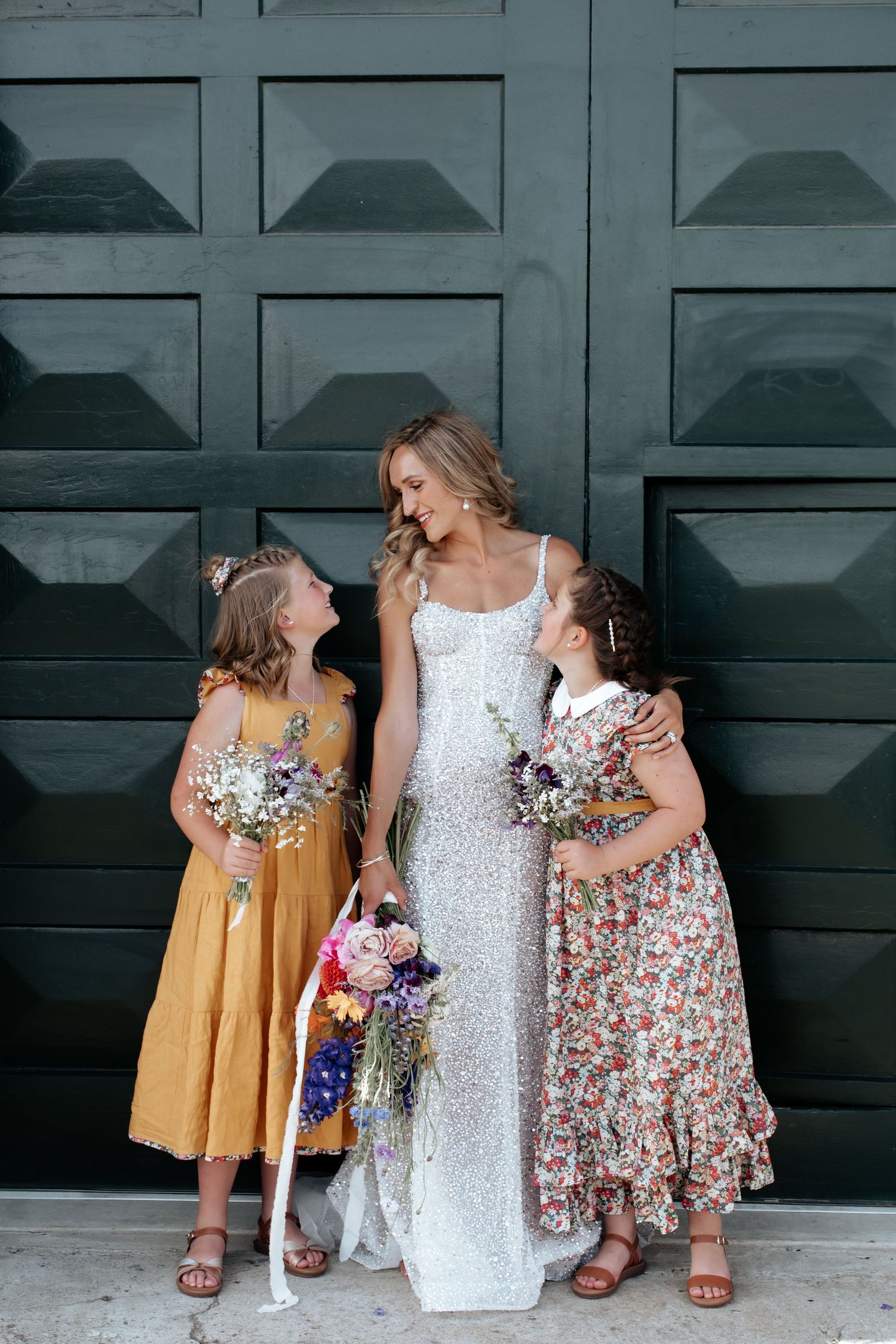 A bride in a sequined gown shares a joyful moment with two young attendants in floral and yellow dresses.