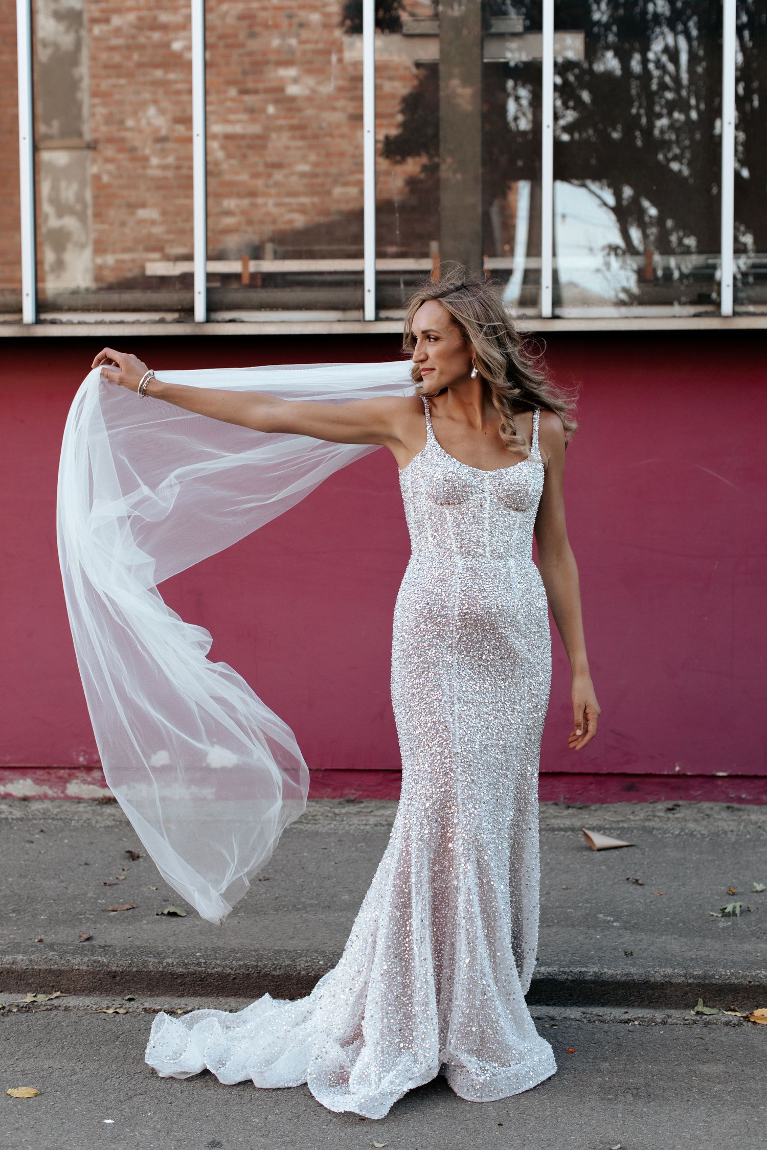 Bride in a fitted sequin dress poses with flowing veil against a red wall background.
