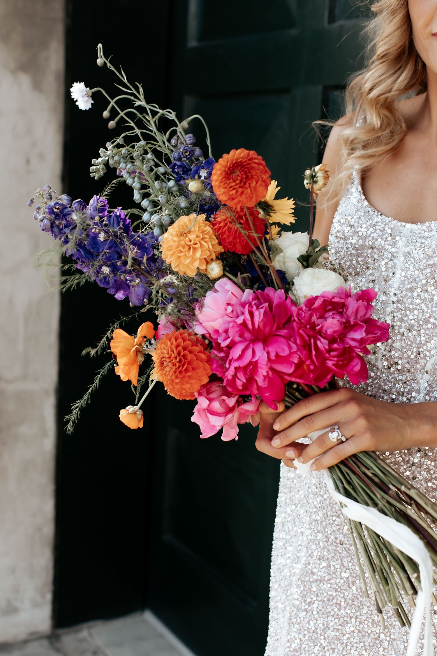 A vibrant wedding bouquet featuring orange, pink, and purple flowers held against a sparkly white dress.