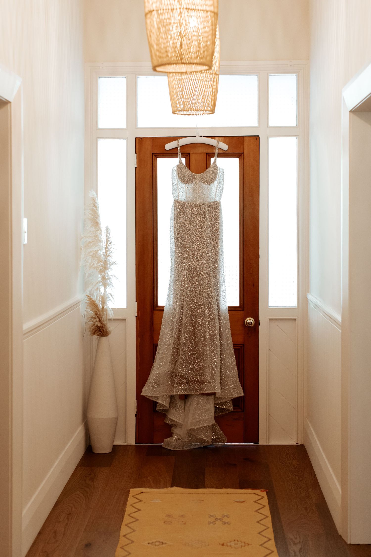 Elegant sequined wedding dress hanging on wooden door in hallway with woven pendant light.