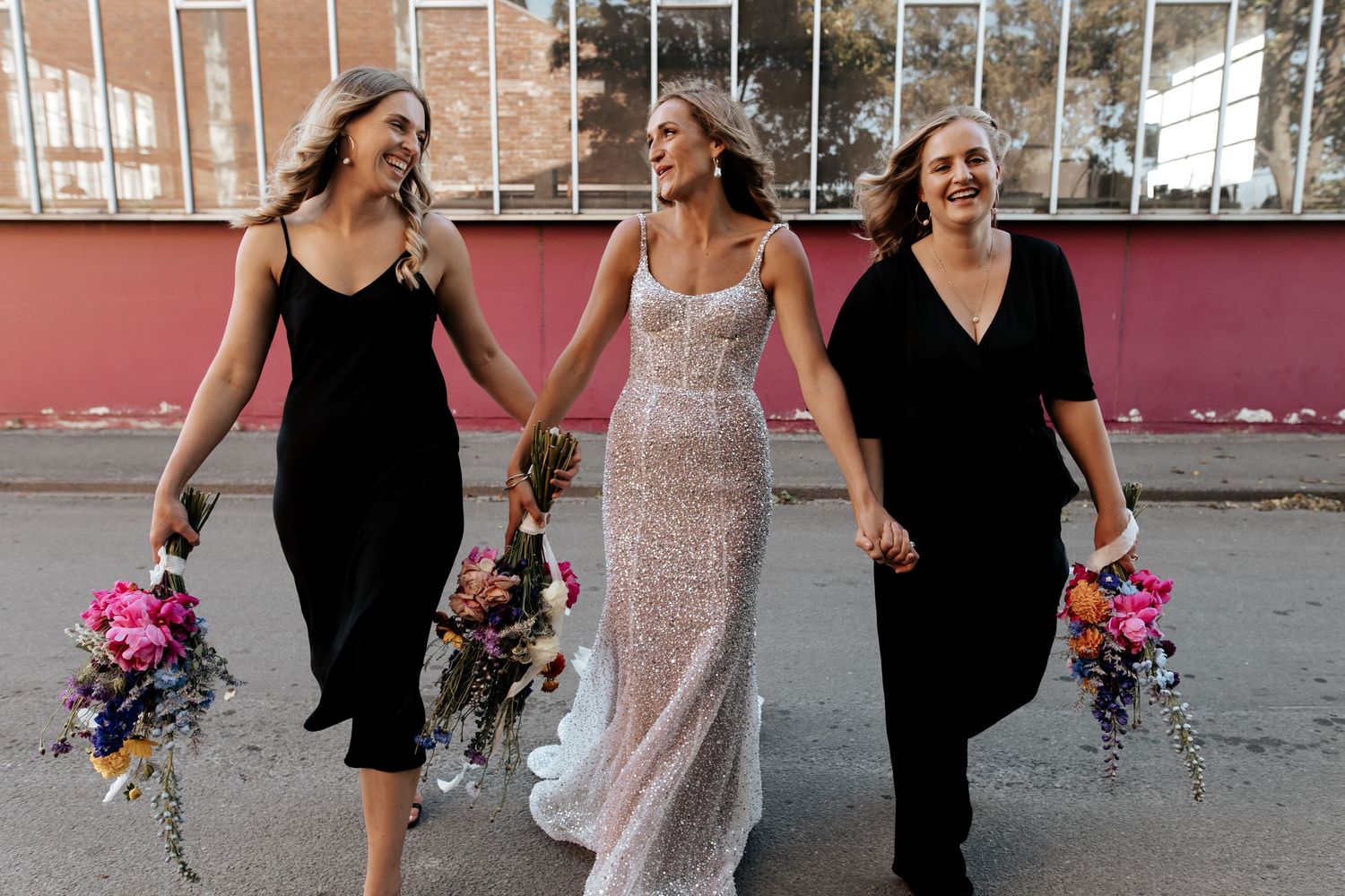 Three women walk together holding colorful bouquets, with the middle one wearing a sparkly white dress.