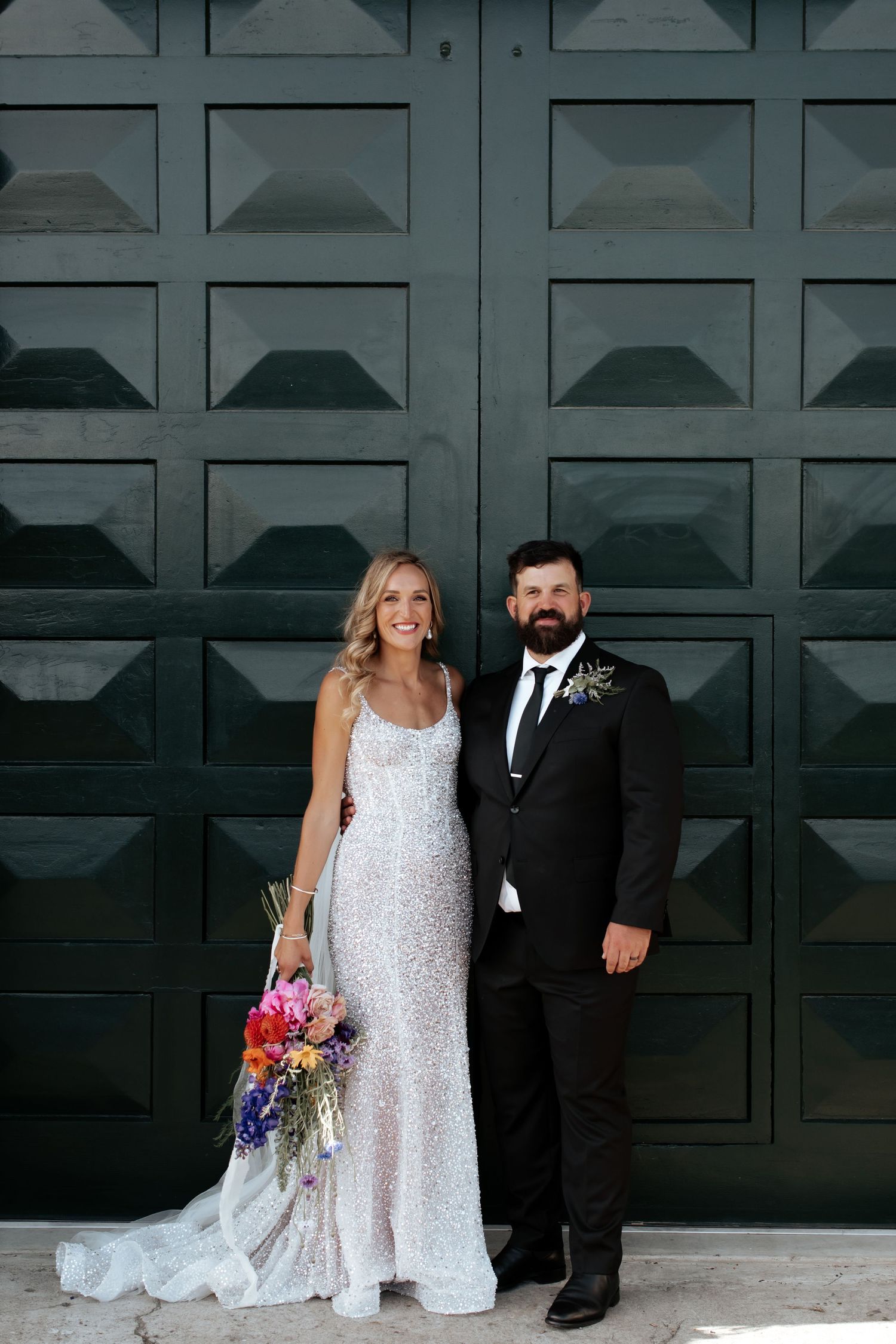 A wedding couple poses formally against a dark geometric door backdrop.
