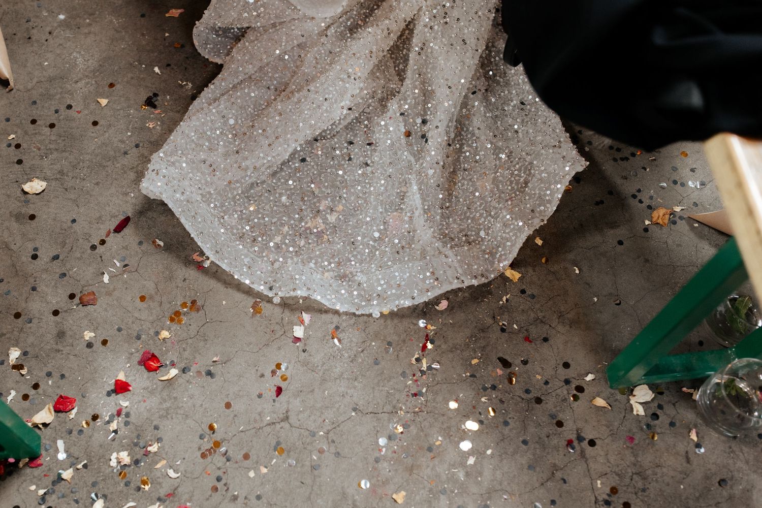 Close-up of sparkly wedding dress hem with scattered confetti on concrete floor.