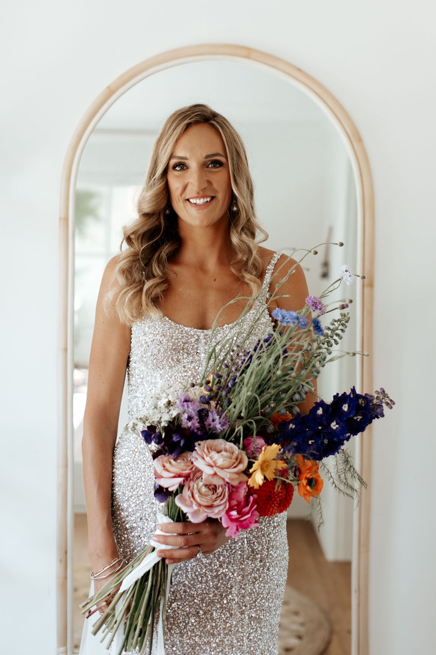 Arched mirror reflection showing sequined wedding dress and vibrant floral bouquet.