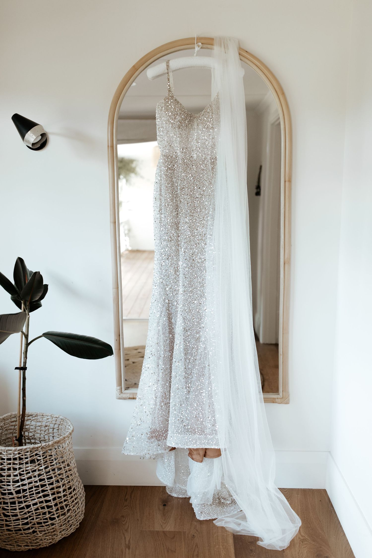 Sparkly white wedding dress hanging on arched mirror with potted plant and woven basket.