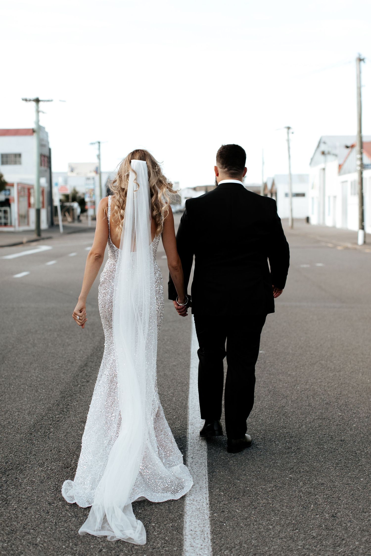 Rear view of newlyweds walking down empty street with bride's veil flowing behind.
