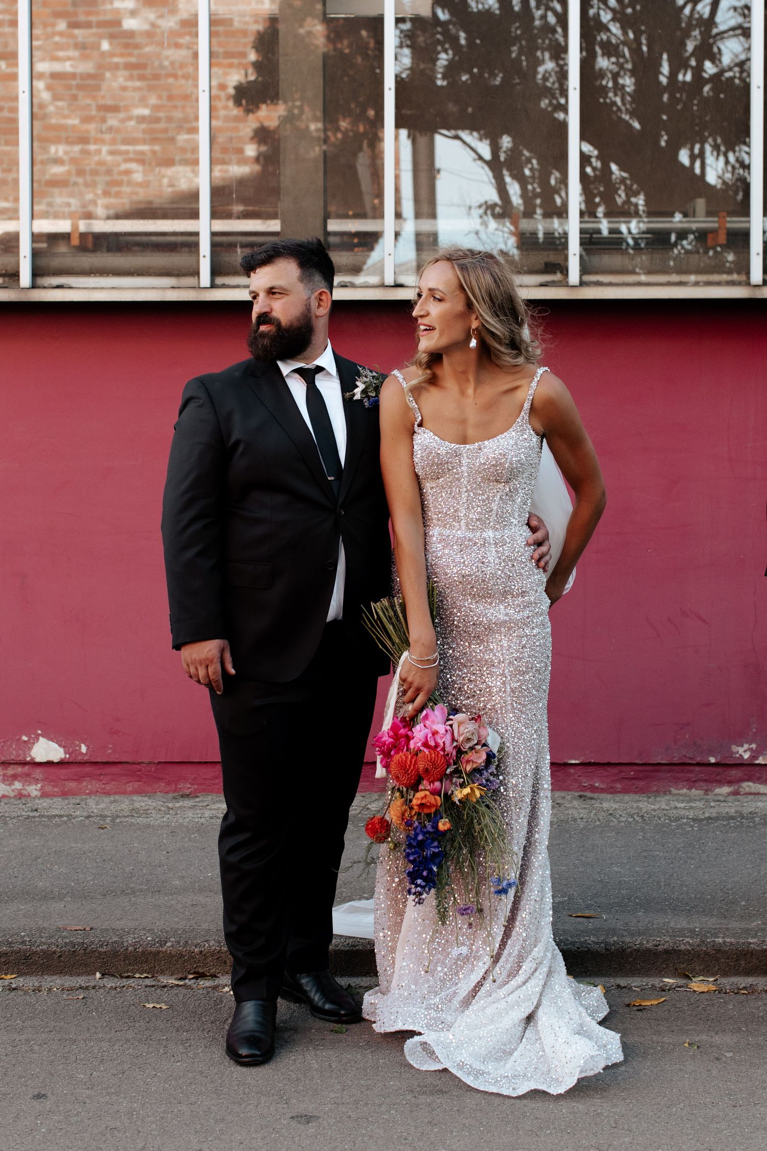 A couple poses against a red wall, with the bride wearing a sparkly white gown and holding a vibrant bouquet.