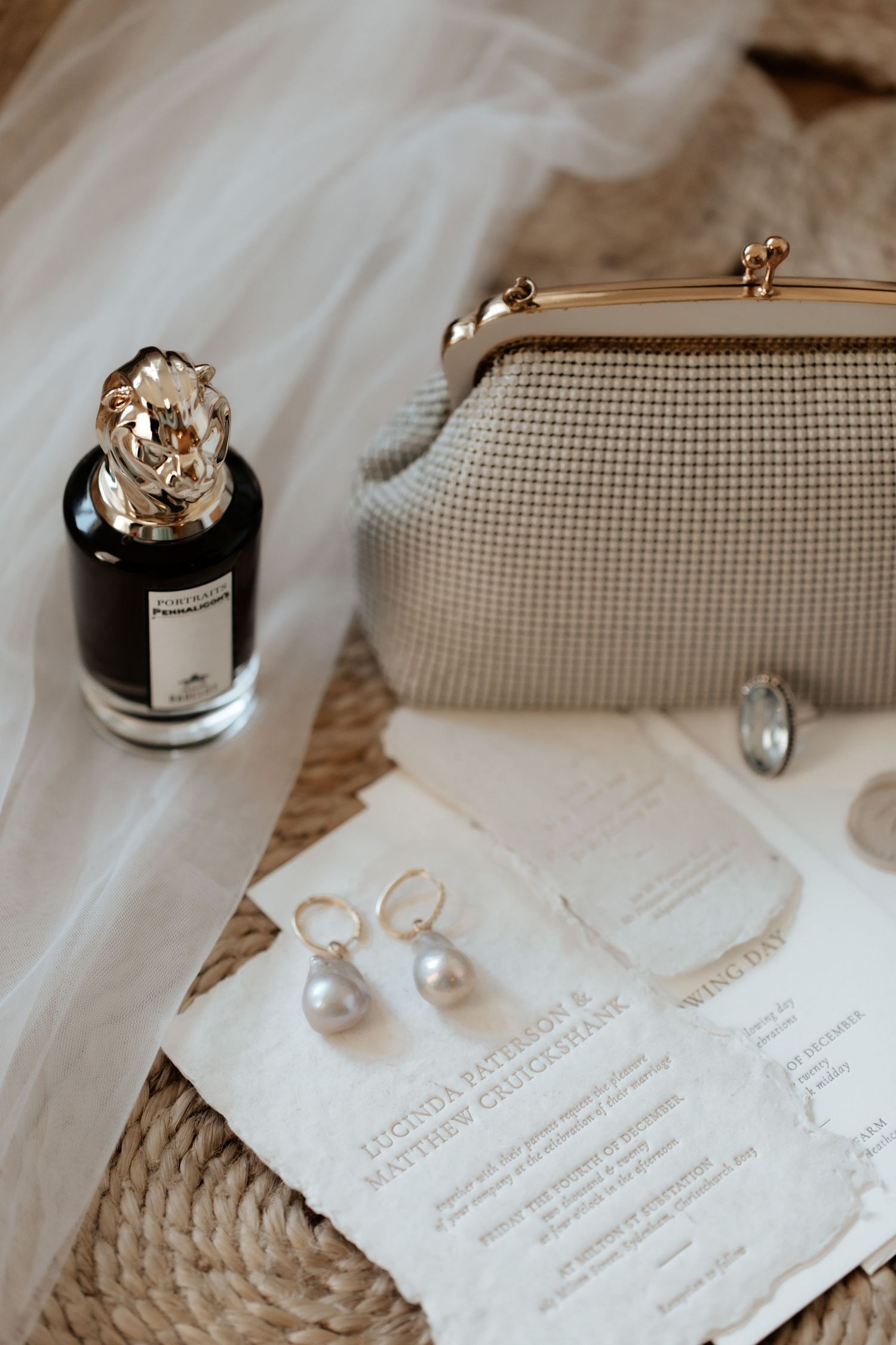 Wedding details including perfume bottle, pearl earrings, white clutch and invitation laid on natural woven surface.