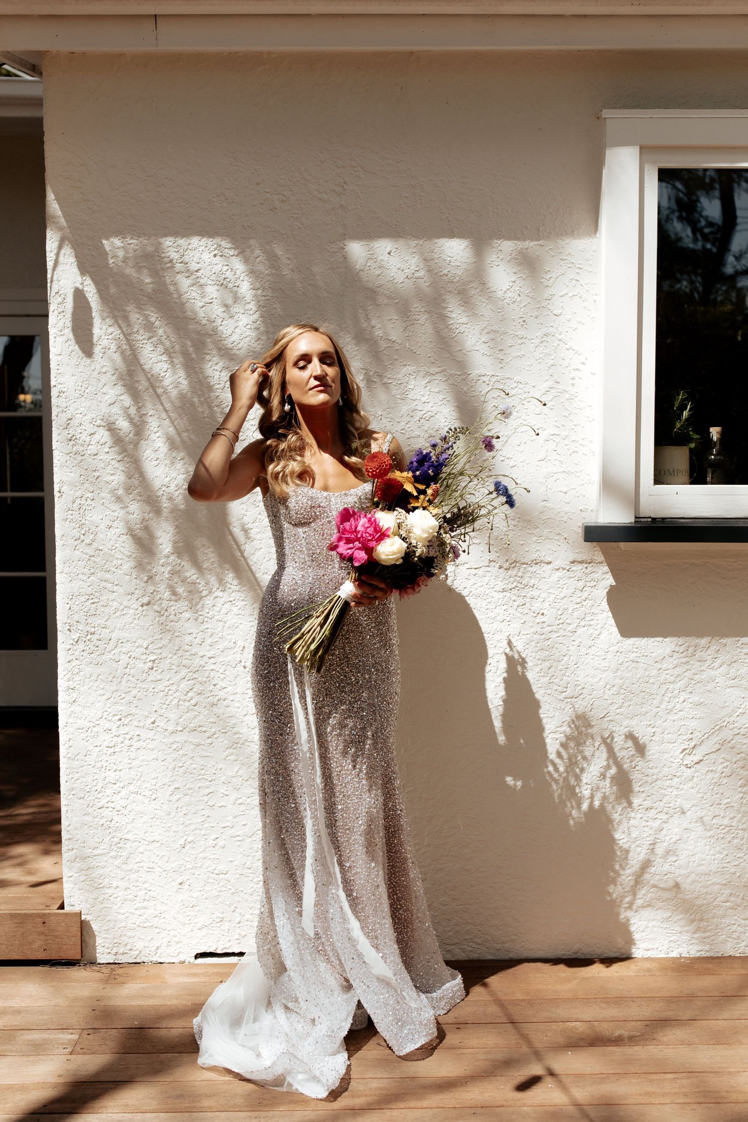 Bride in sequined gown holding vibrant bouquet poses against white wall in sunlight.